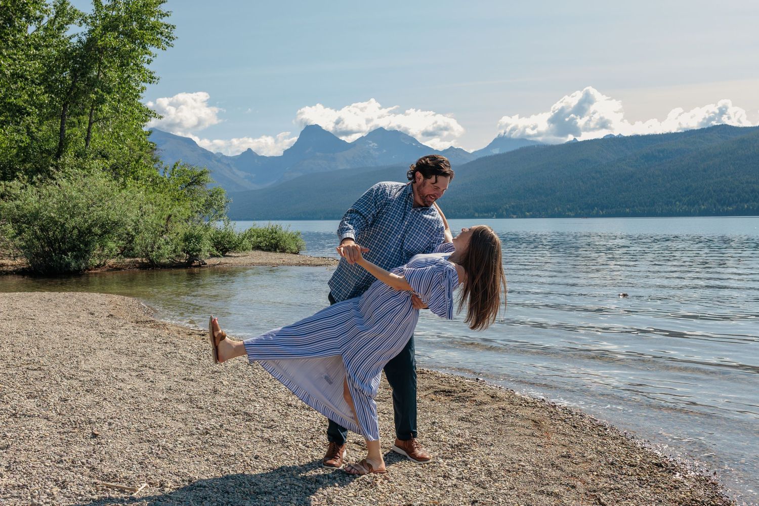 A couple dances on a beach shoreline with scenic mountain views and lake waters in the background.