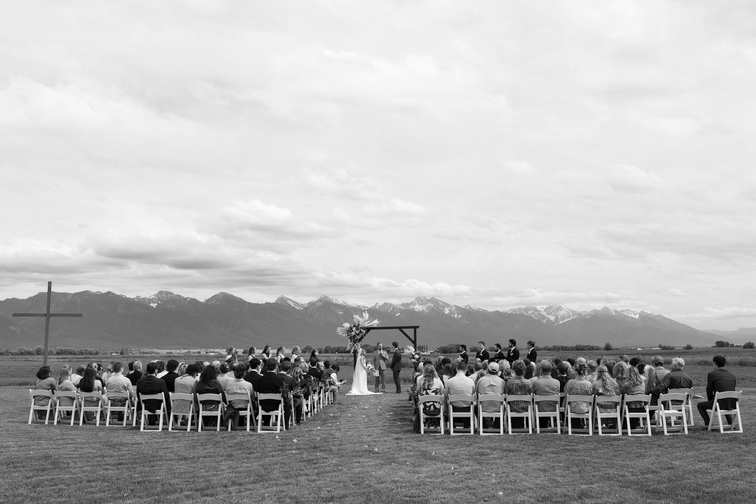 Montana Outdoor wedding ceremony with mission mountain backdrop, guests seated in white chairs facing couple at wooden arch.