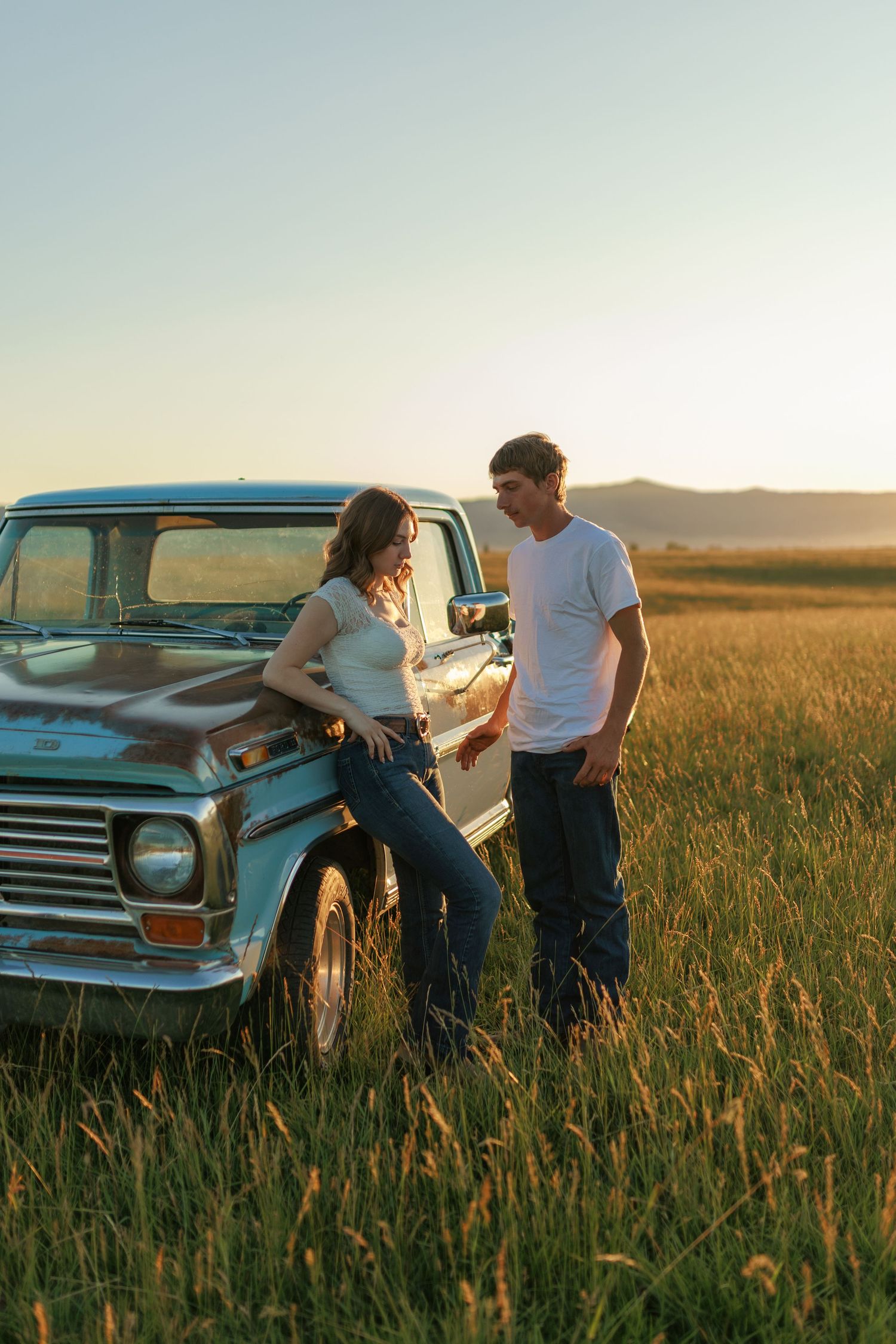 Couple in casual denim poses by vintage blue pickup truck in sunny wheat field.