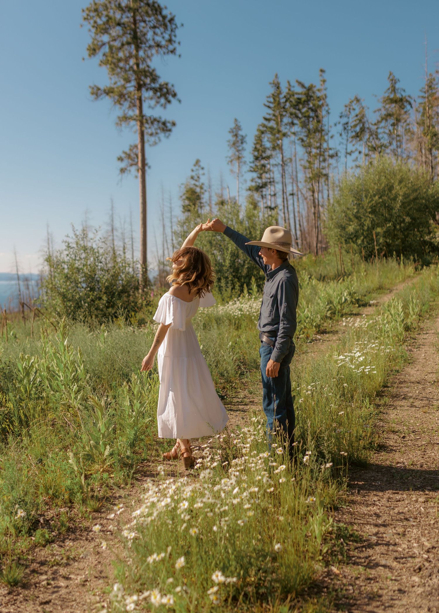 A couple in casual attire strolls along a scenic mountain path surrounded by white daisies.