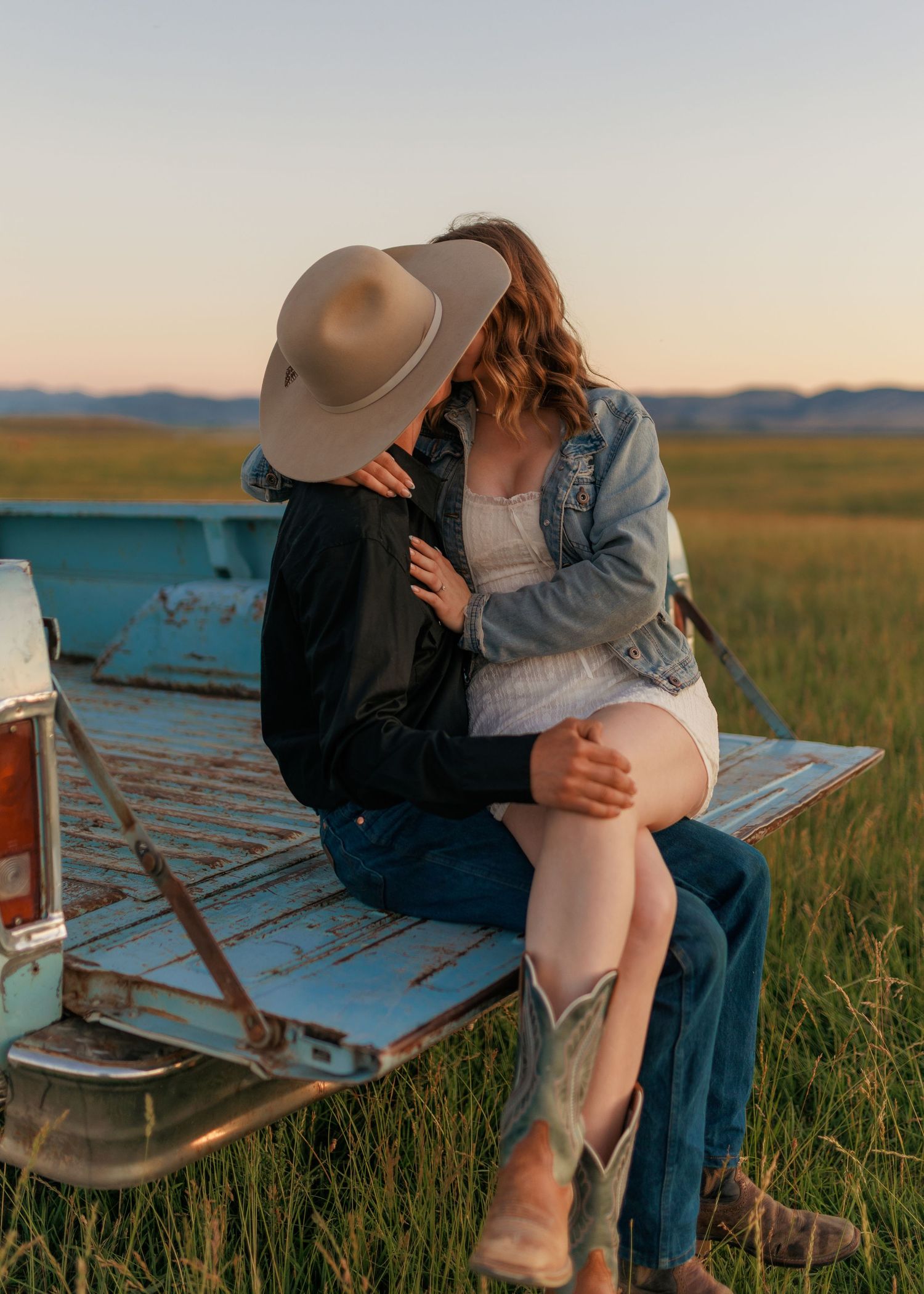 Couple sitting on tailgate of vintage truck in prairie field at sunset.