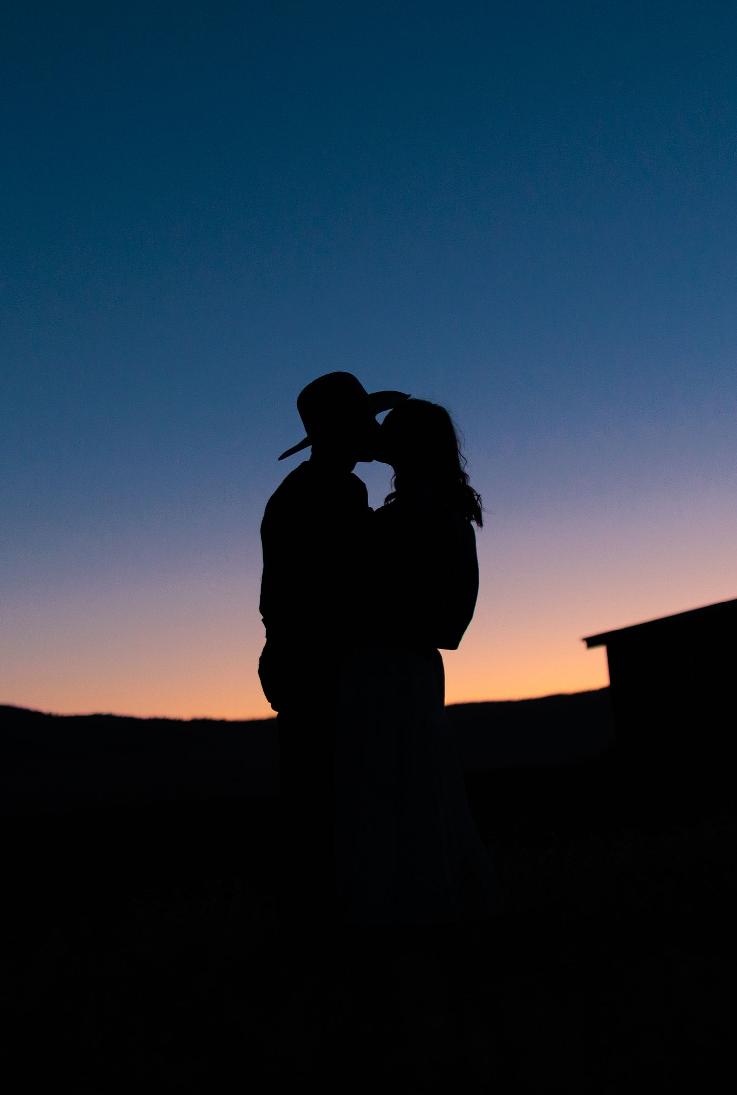 Silhouette of a couple embracing against a sunset sky with a barn in the background.