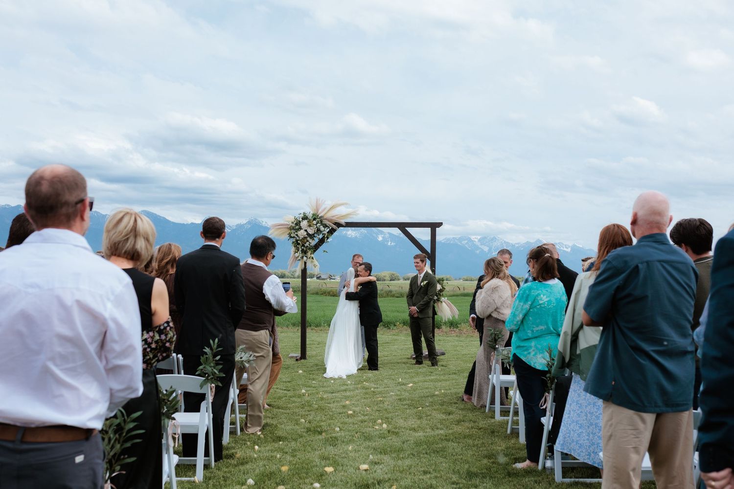 Montana Outdoor wedding ceremony with mission mountain backdrop, guests seated in white chairs facing couple at wooden arch.