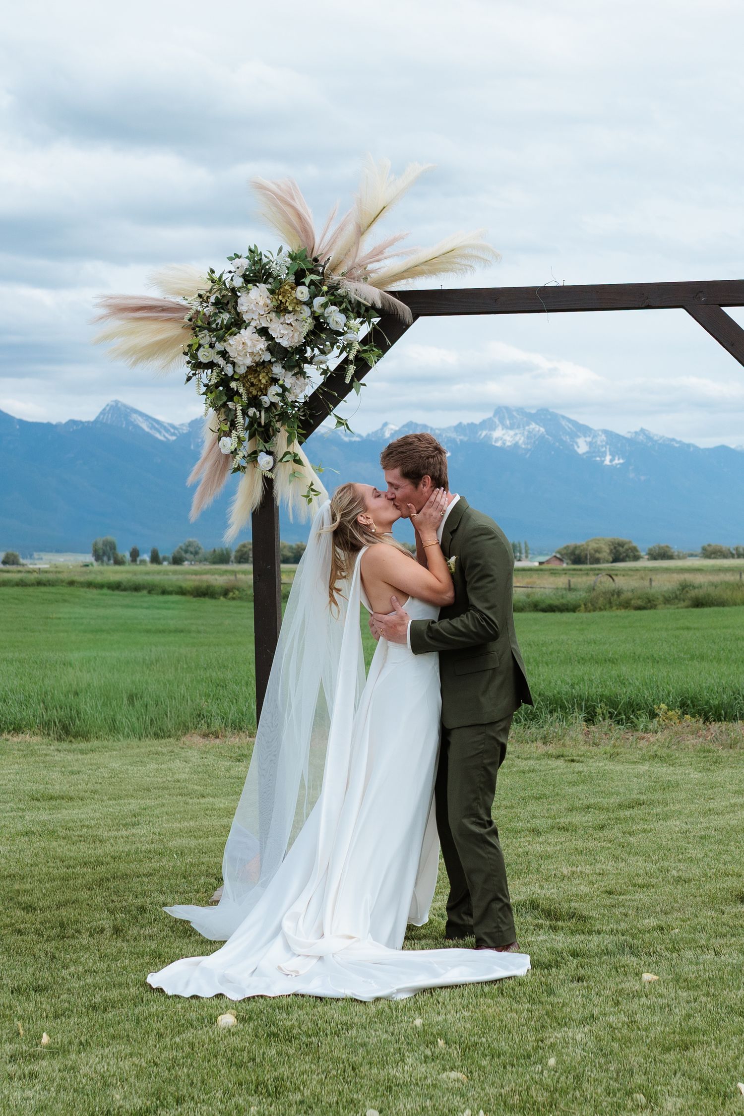 Bride and groom share first kiss under rustic wooden arch with pampas grass florals, Montana mountains backdrop.