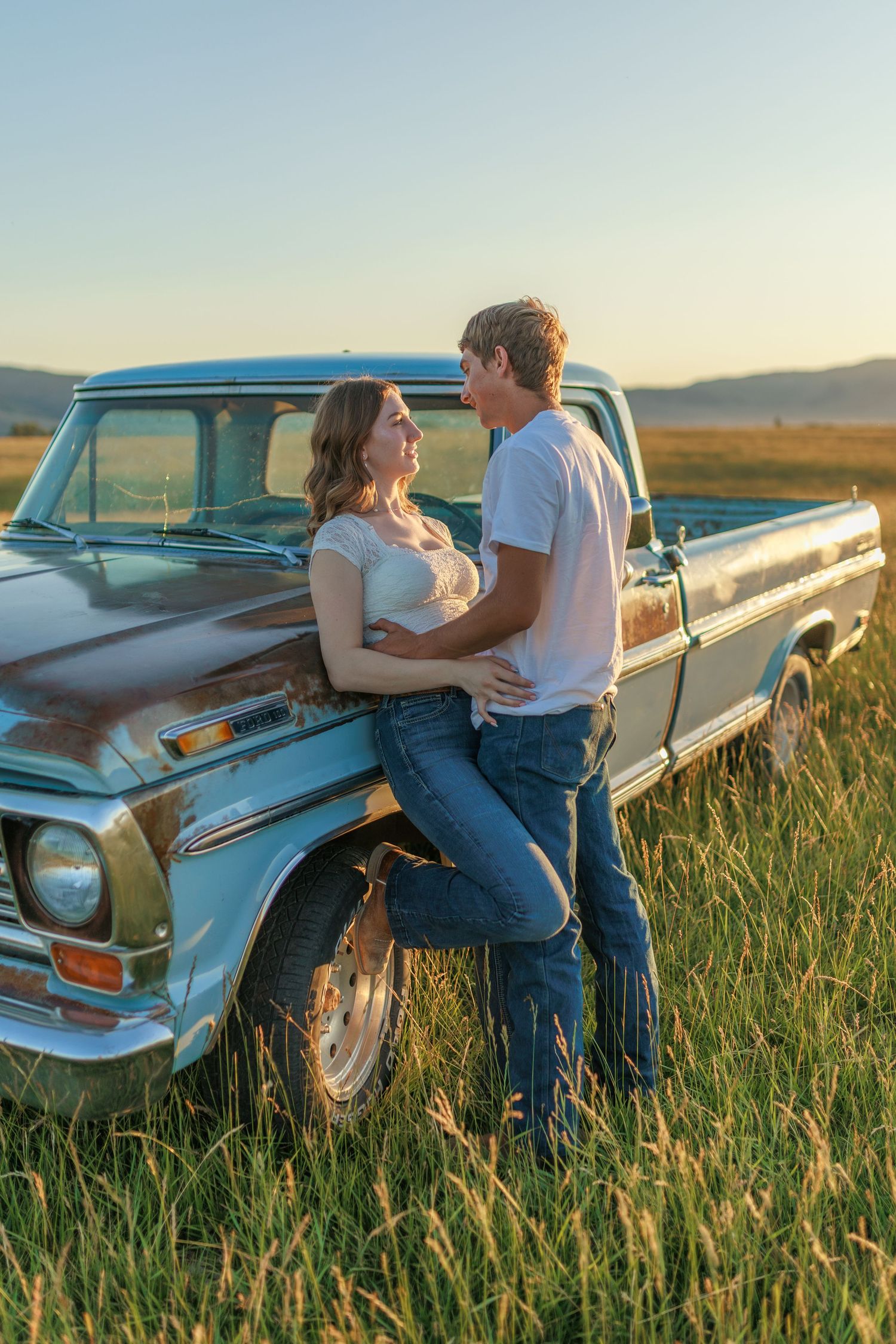 Couple poses against vintage blue pickup truck in field of tall grass at sunset.