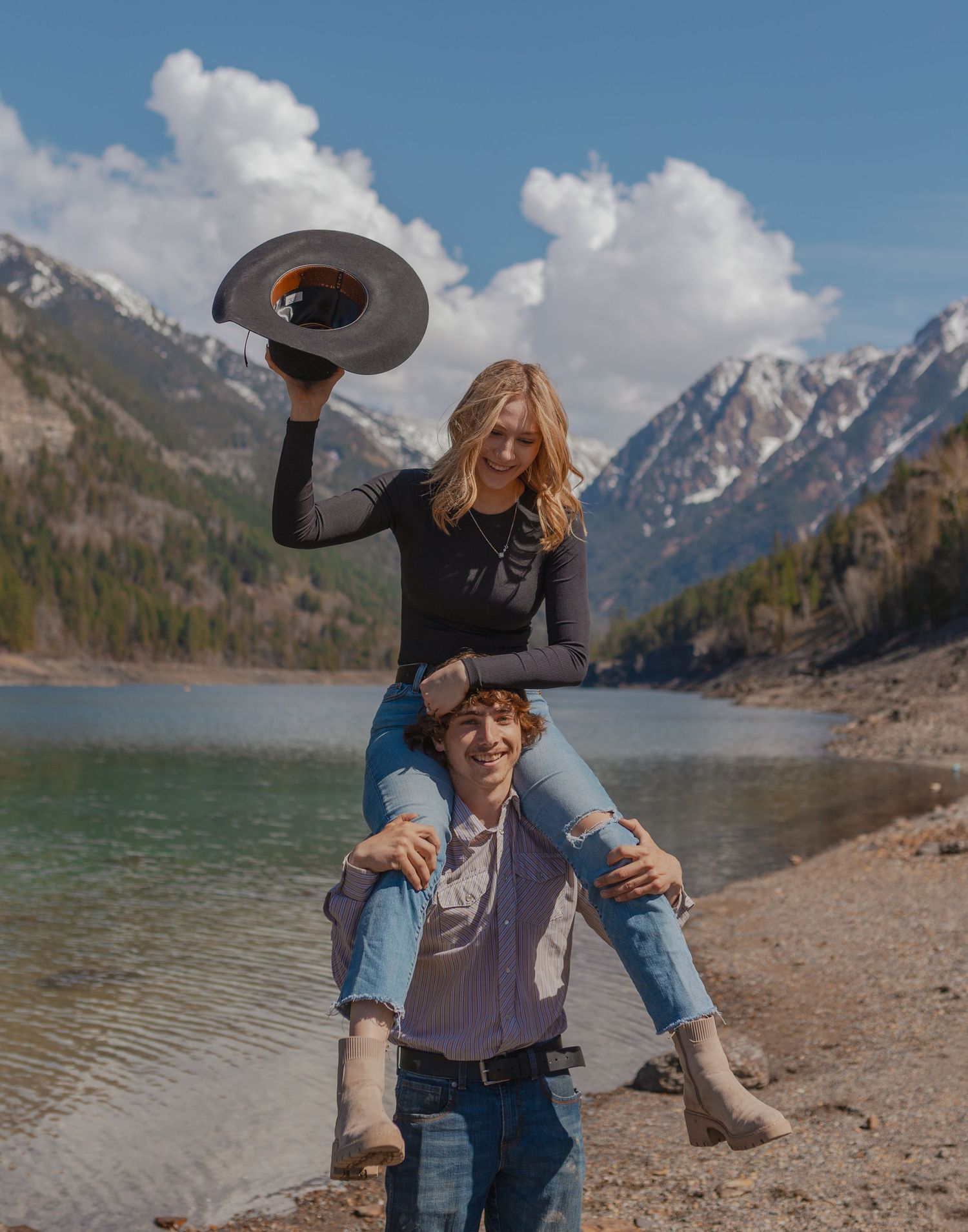 A playful couple has fun with a cowboy hat by a scenic mountain lake.