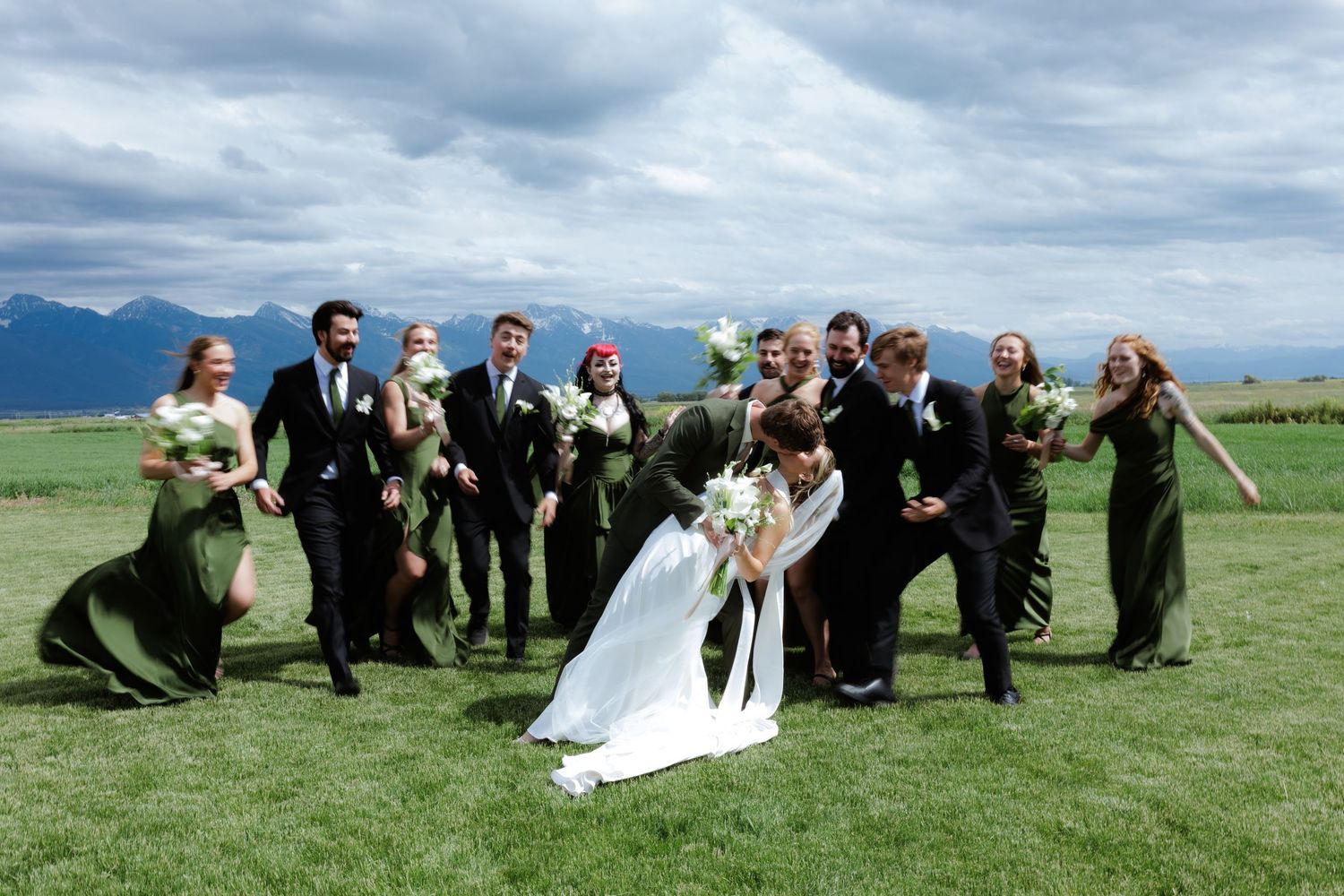 Wedding party photo with bride and groom kissing while bridal party celebrate outdoors with Mission mountains in background.