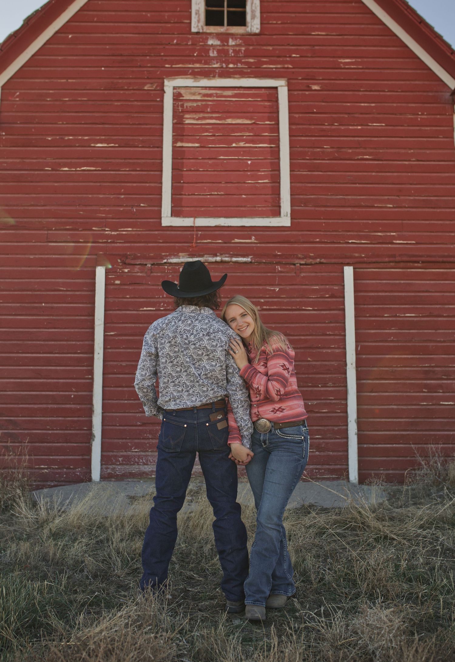 A couple shares an intimate moment while standing in front of a weathered red barn.
