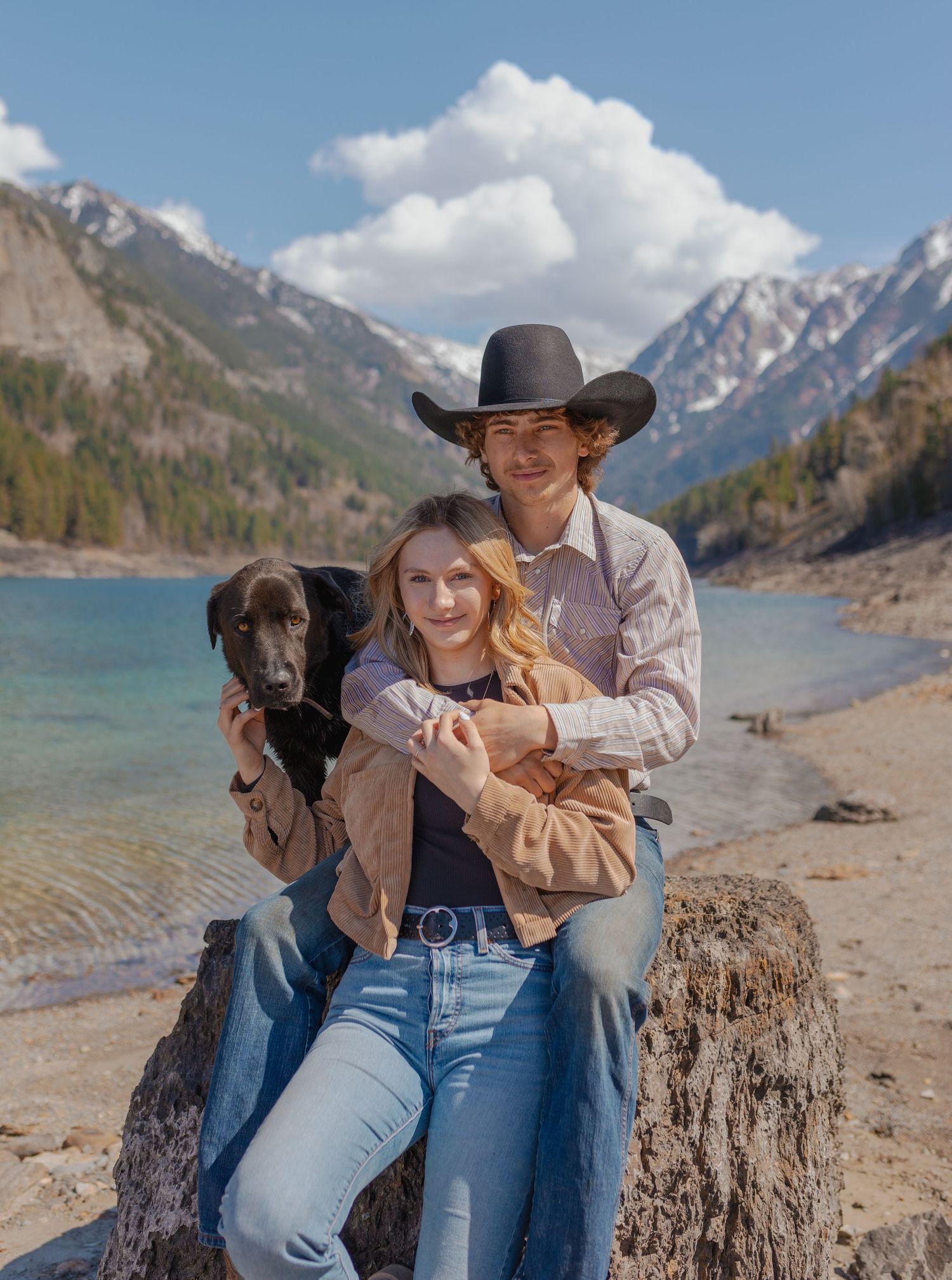 A couple and their black dog pose together on a rock by a mountain lake.