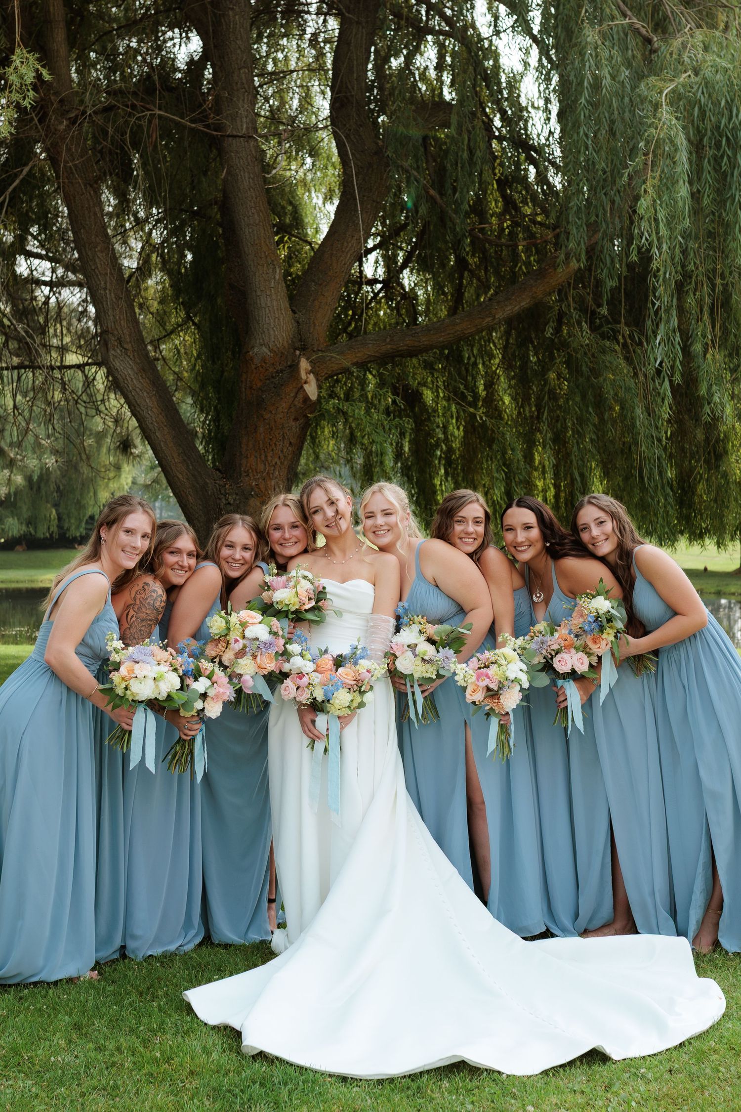 Bride in white gown poses with eight bridesmaids in dusty blue dresses holding colorful bouquets under a willow tree in Montana.