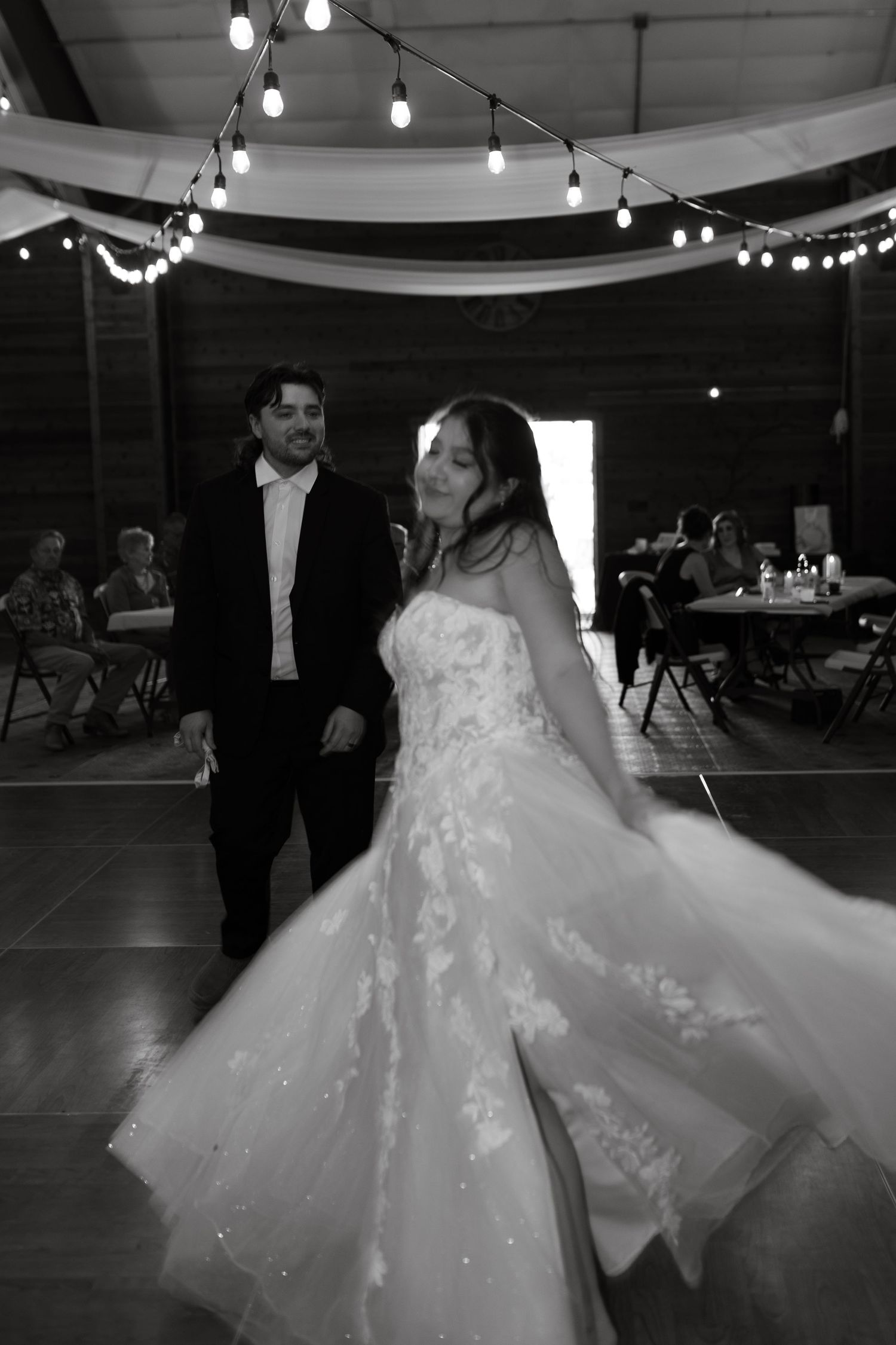 Black and white photo of bride in lace gown twirling with groom in barn venue adorned with string lights.