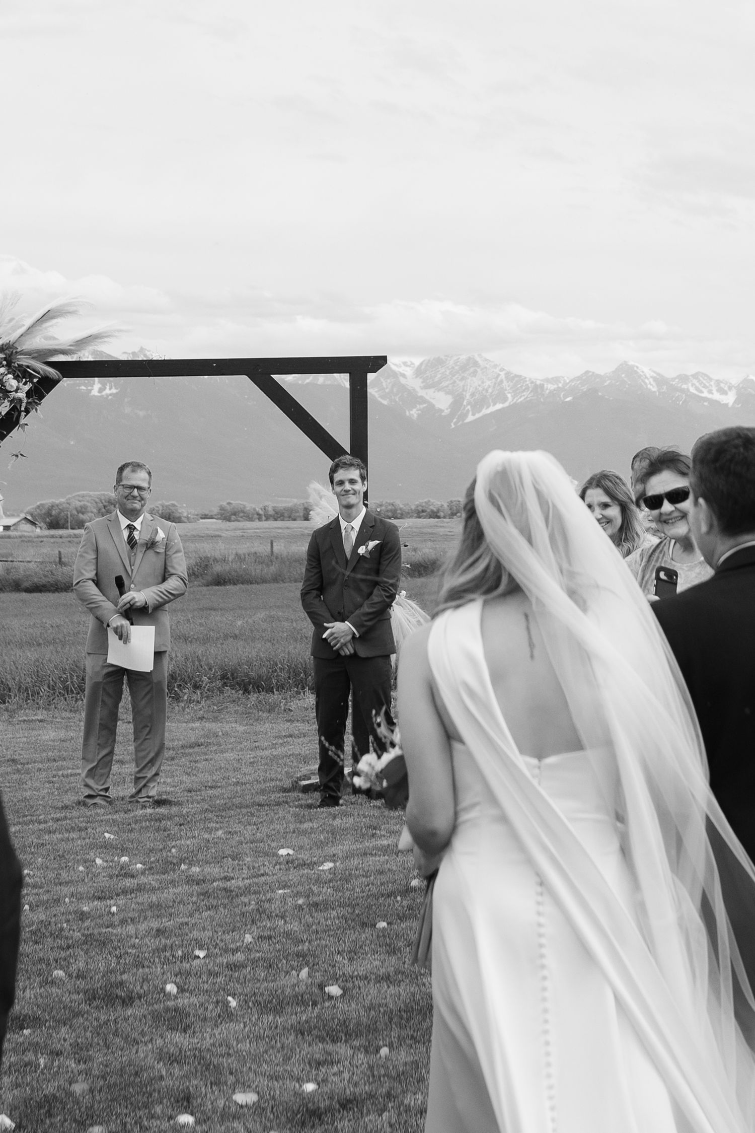 Black and white outdoor wedding ceremony as bride walks with dad toward emotional groom at rustic arch, mission mountains in background.