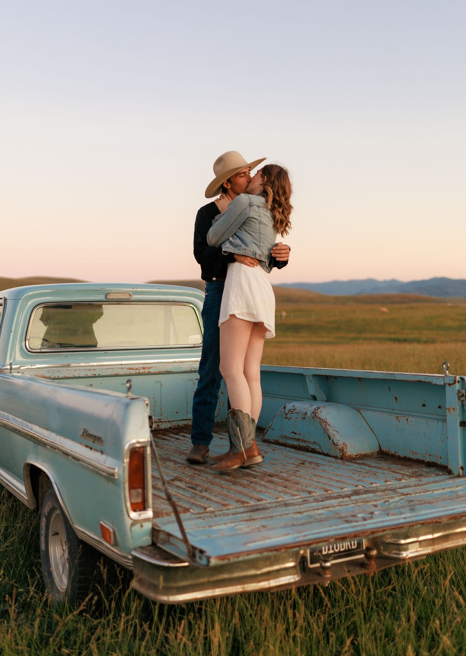 Vintage turquoise pickup truck parked in tall grass with mountains visible in the distance.
