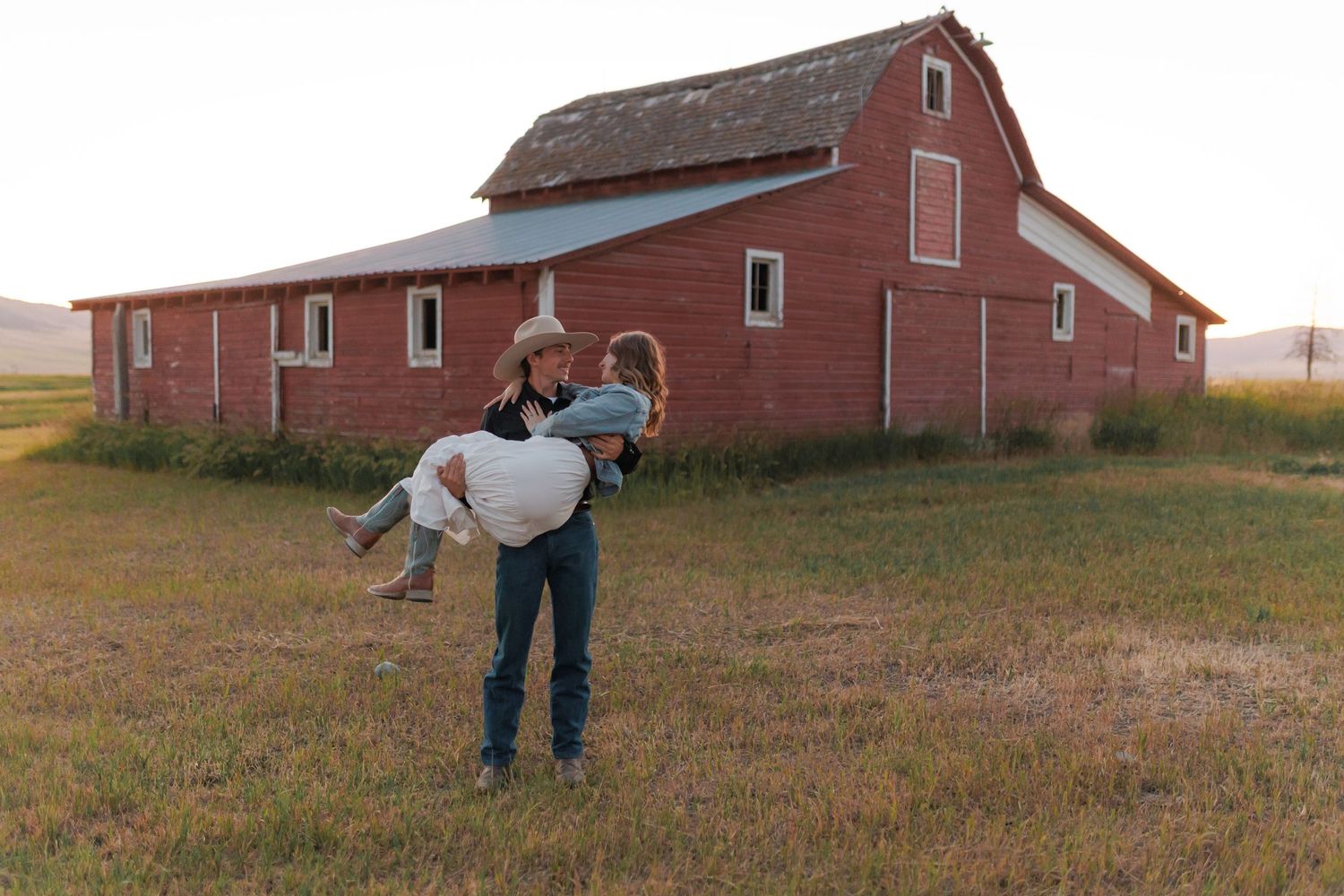 Couple dancing near rustic red barn buildings in countryside setting.