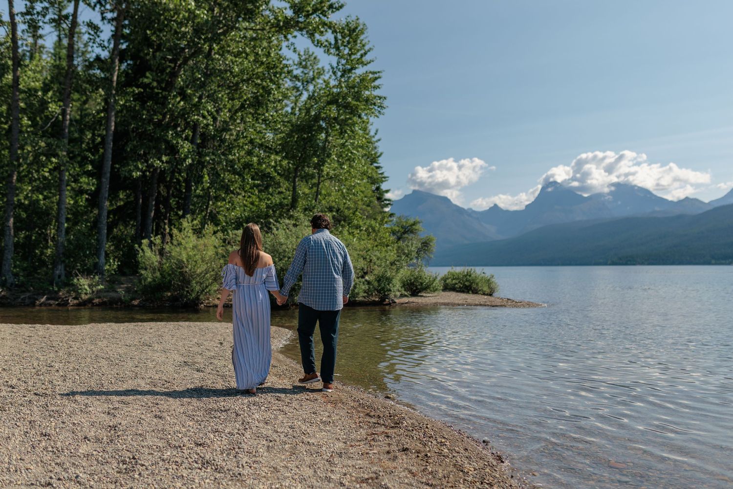 A couple walks along a scenic lakeshore with mountains and evergreen trees in the background on a sunny day.