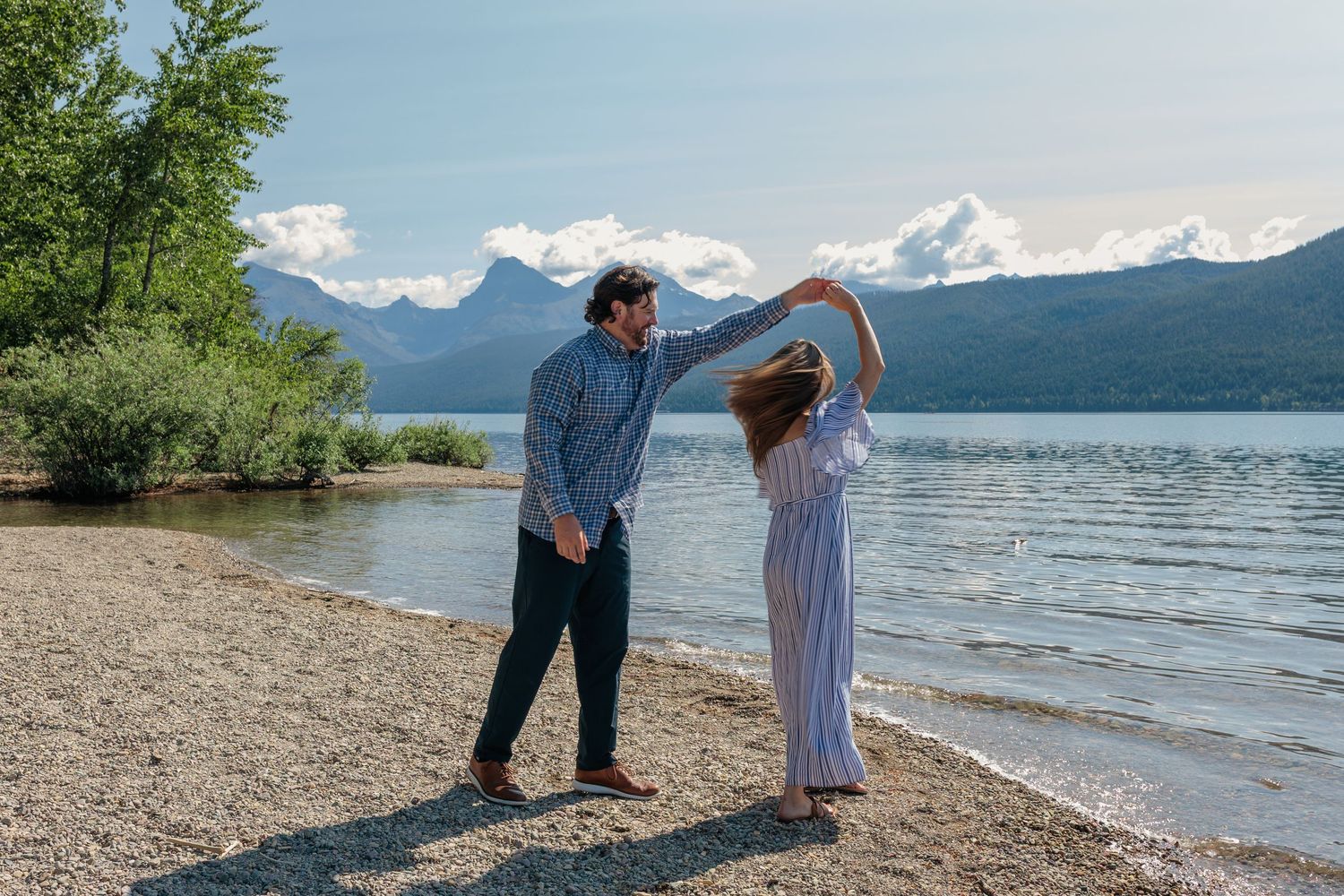 A couple dances playfully on a rocky beach with mountains and a serene lake in the backdrop.