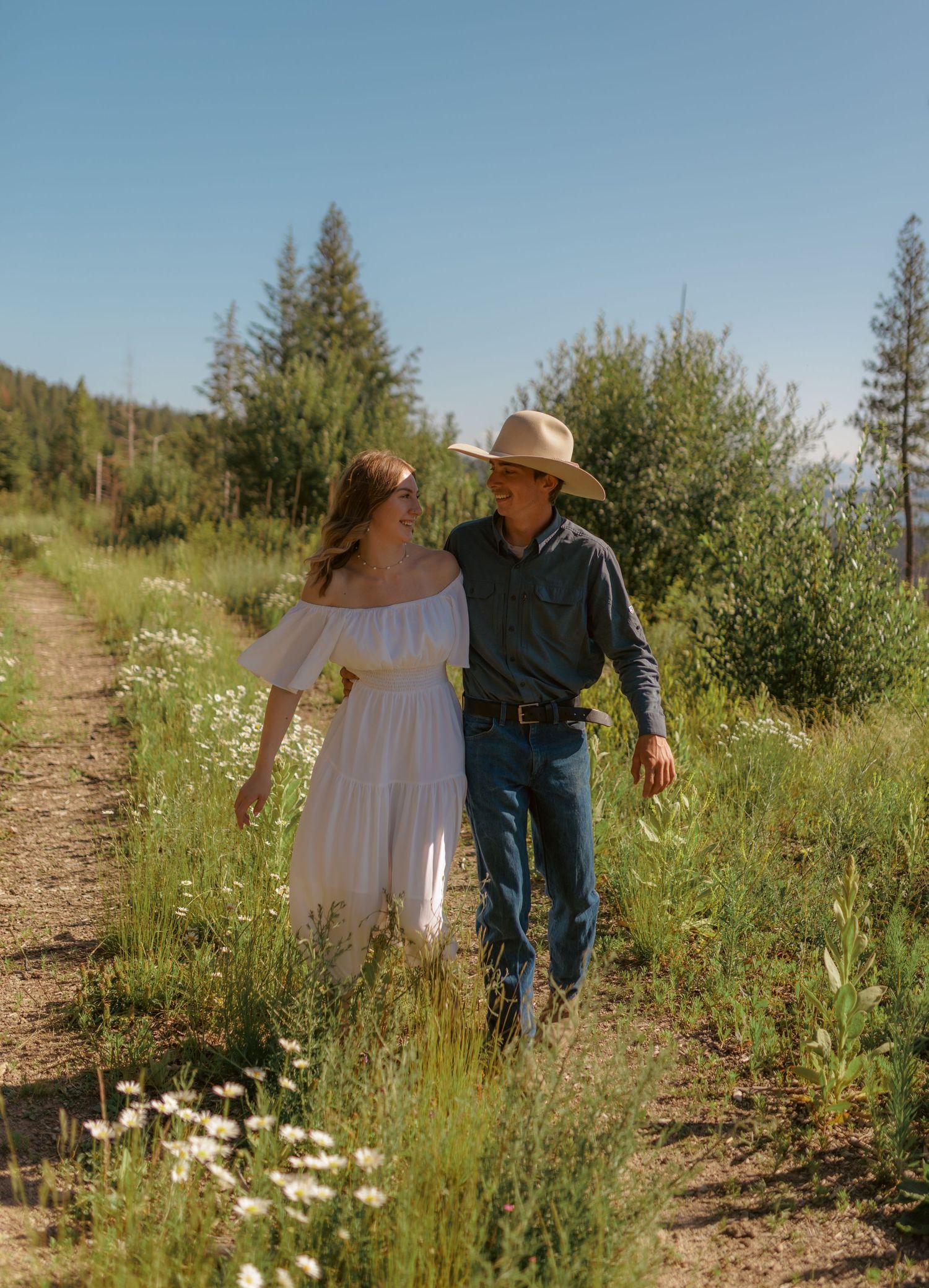 Two people walk hand in hand through a mountain trail dotted with wildflowers.