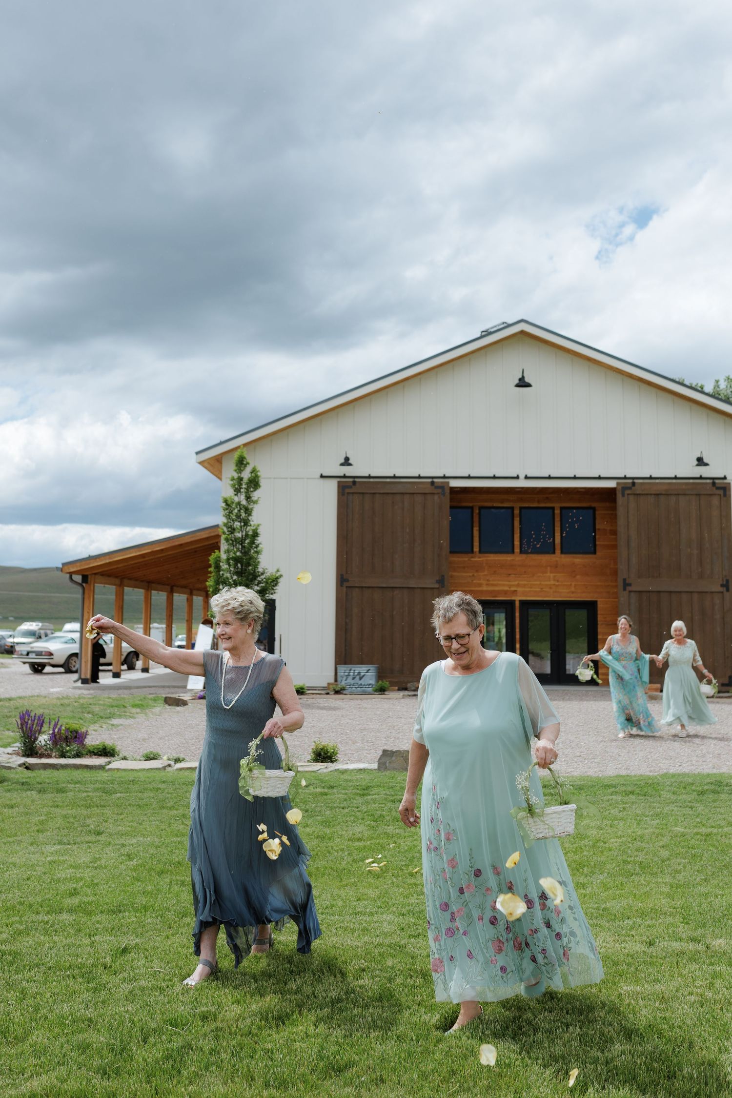 Bride and groom's grandmas scatter flower petals on grass outside a rustic barn wedding venue in Montana with mission mountain views.