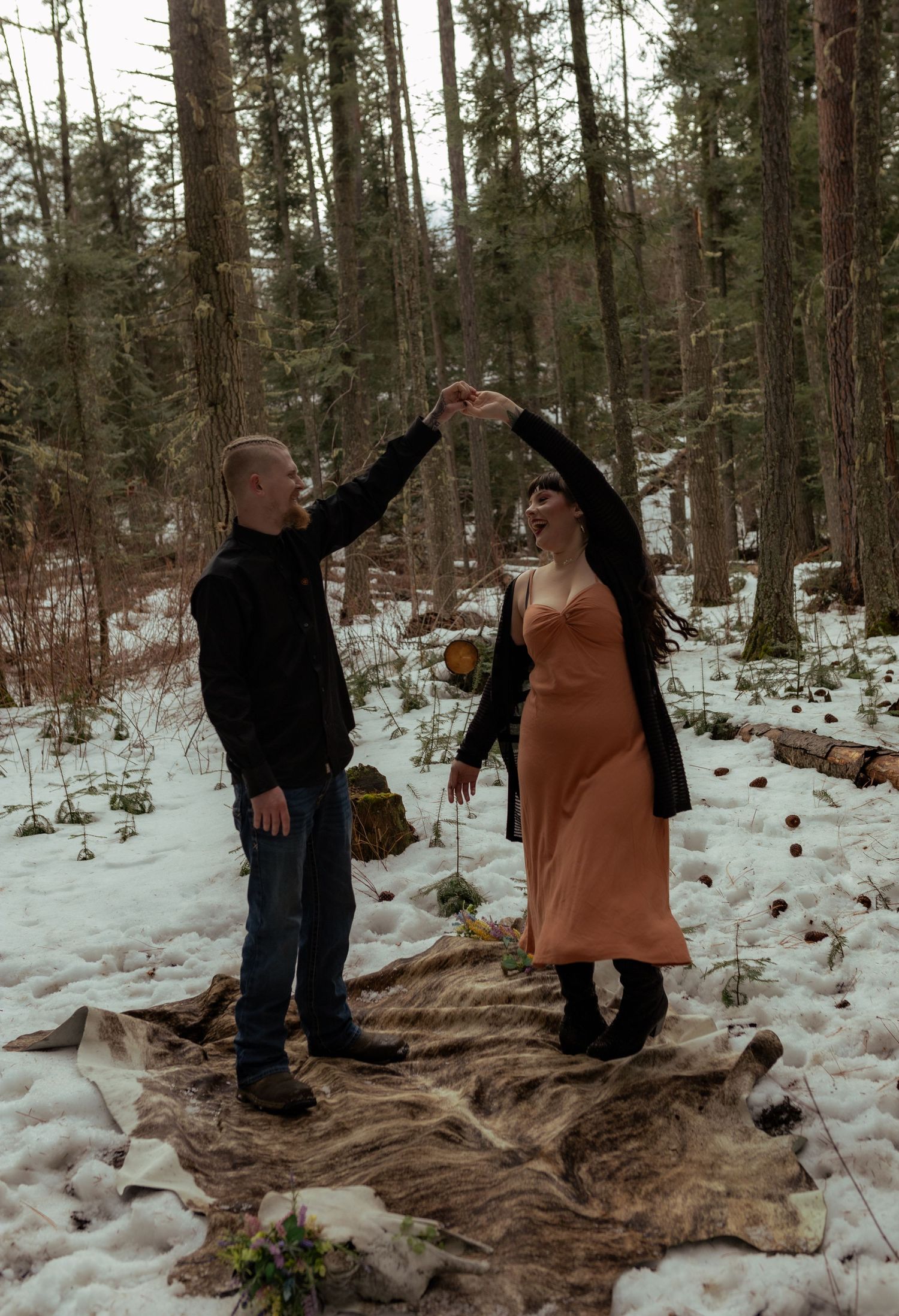 A couple dances on a blanket in a snowy forest setting wearing winter attire.