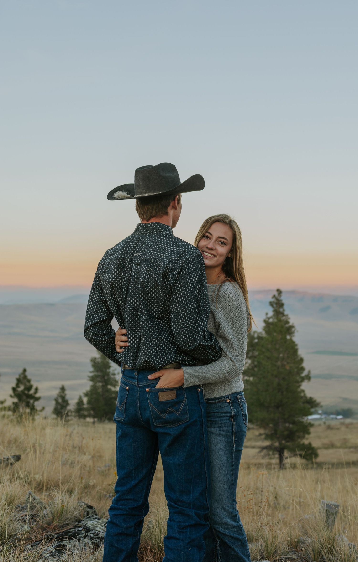 Couple in casual western wear embraces on hilltop overlooking mountain valley at dusk.