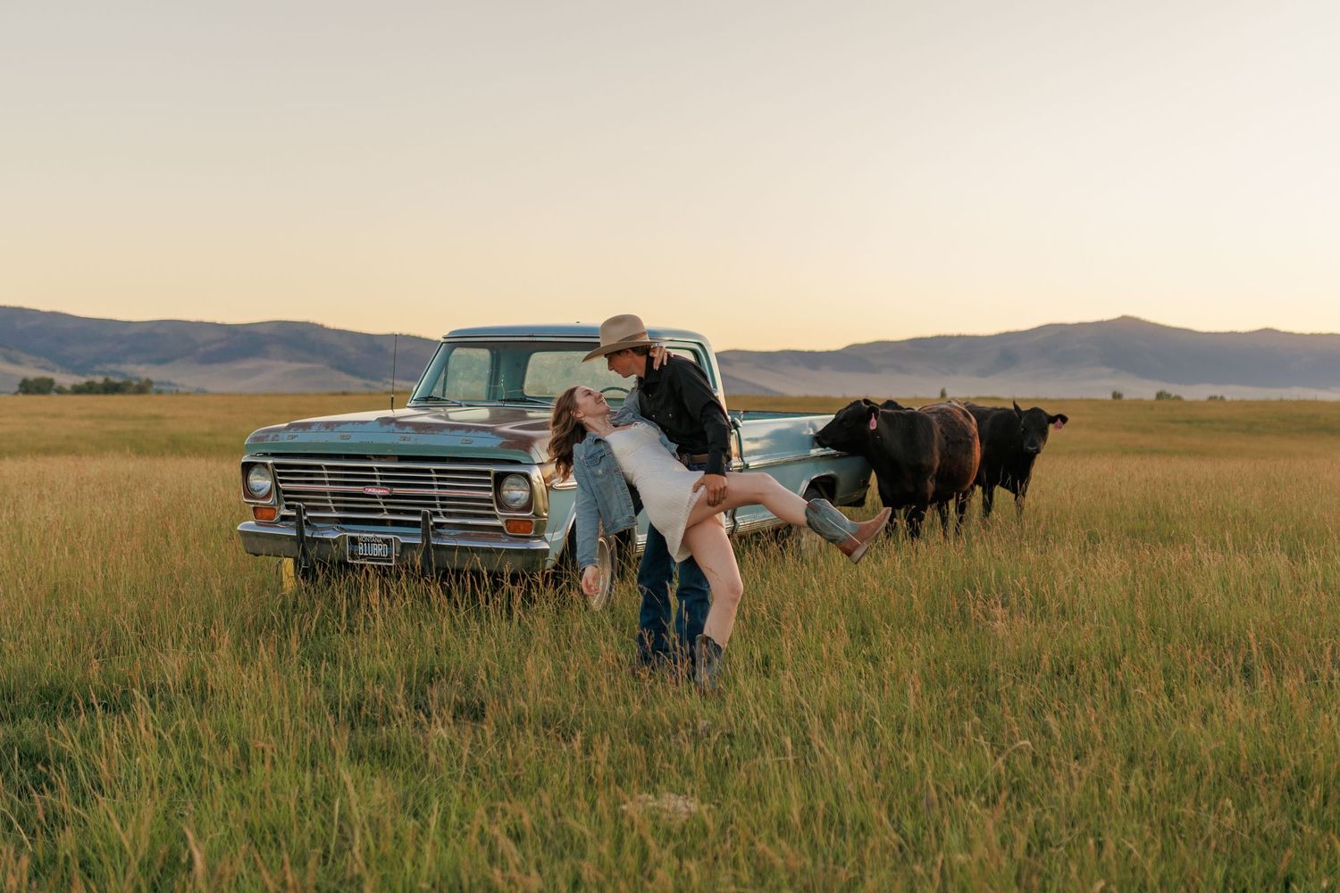 Romantic couple embraces near a vintage car in a golden prairie field at sunset.