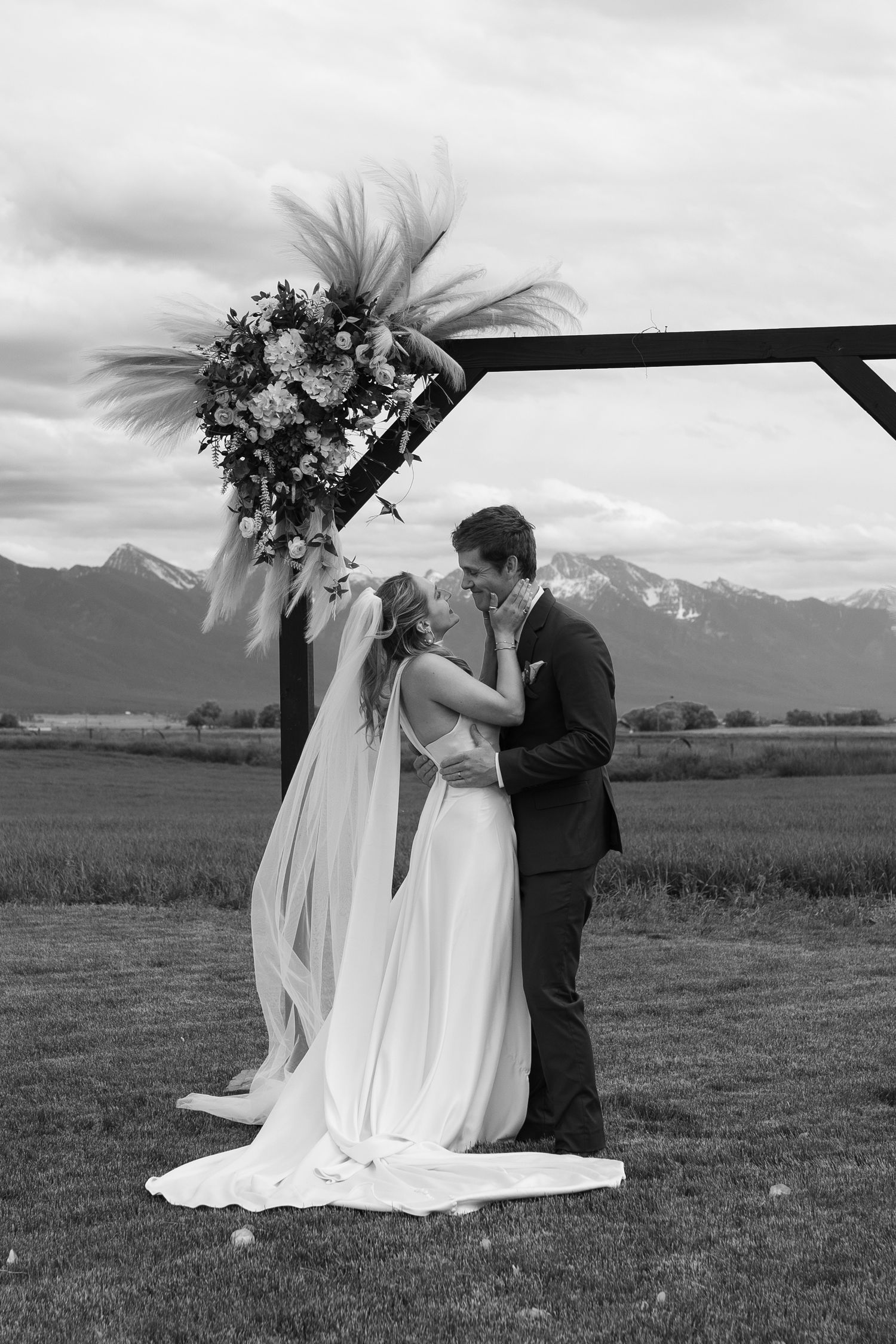 Black and white photo of bride and groom sharing first kiss under floral arch with Montana mission mountain backdrop.