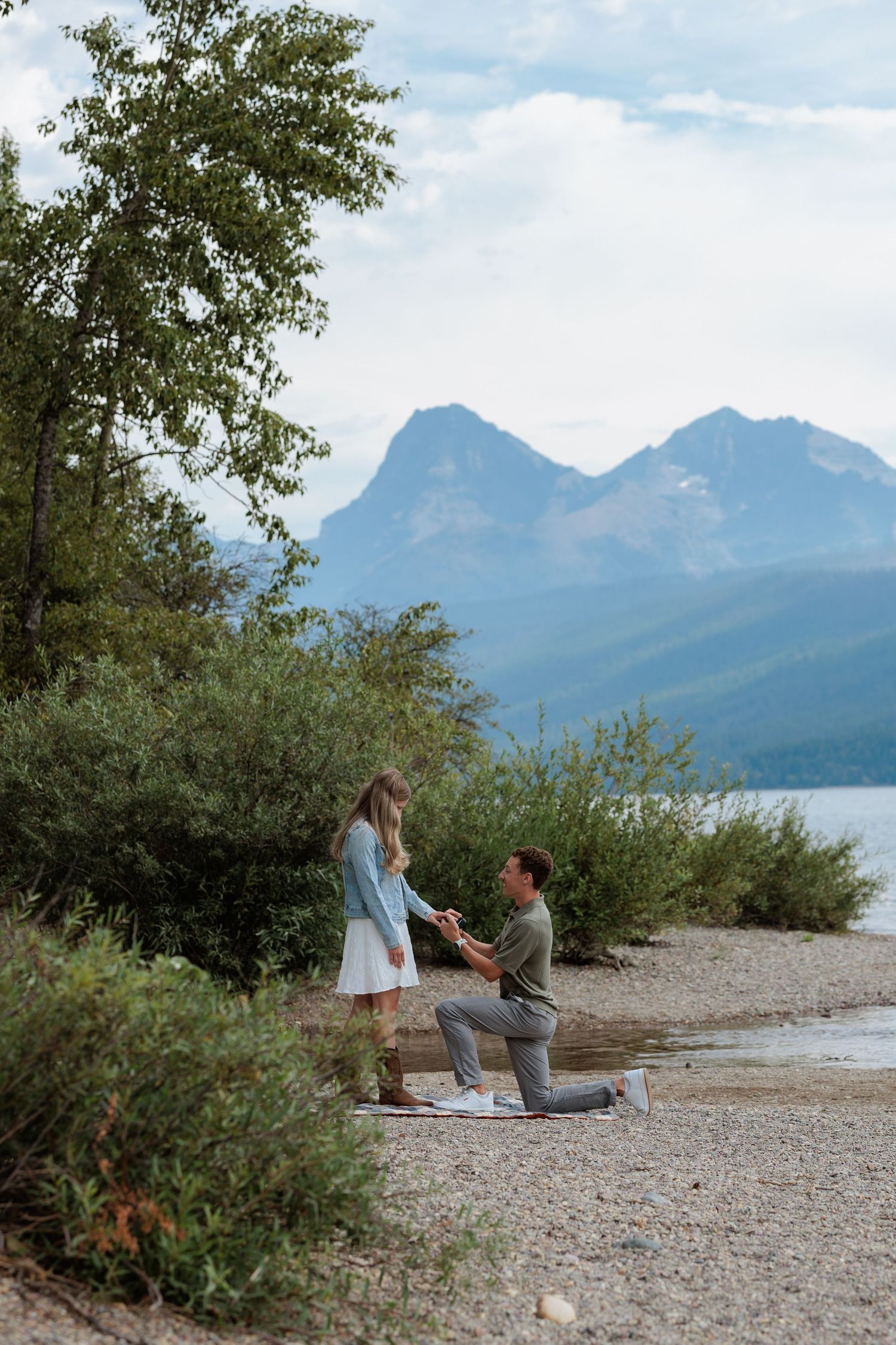 Man proposes on one knee to woman on rocky lakeside beach with stunning mountain backdrop in Glacier National Park at Fish Creek.