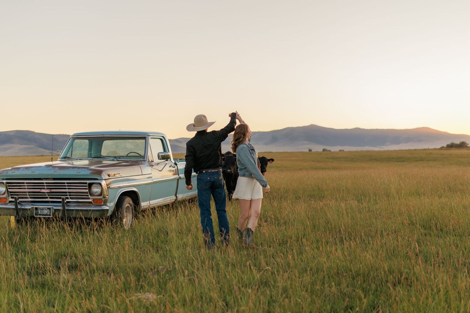 Couple stands by classic Ford truck in vast prairie field at golden hour.