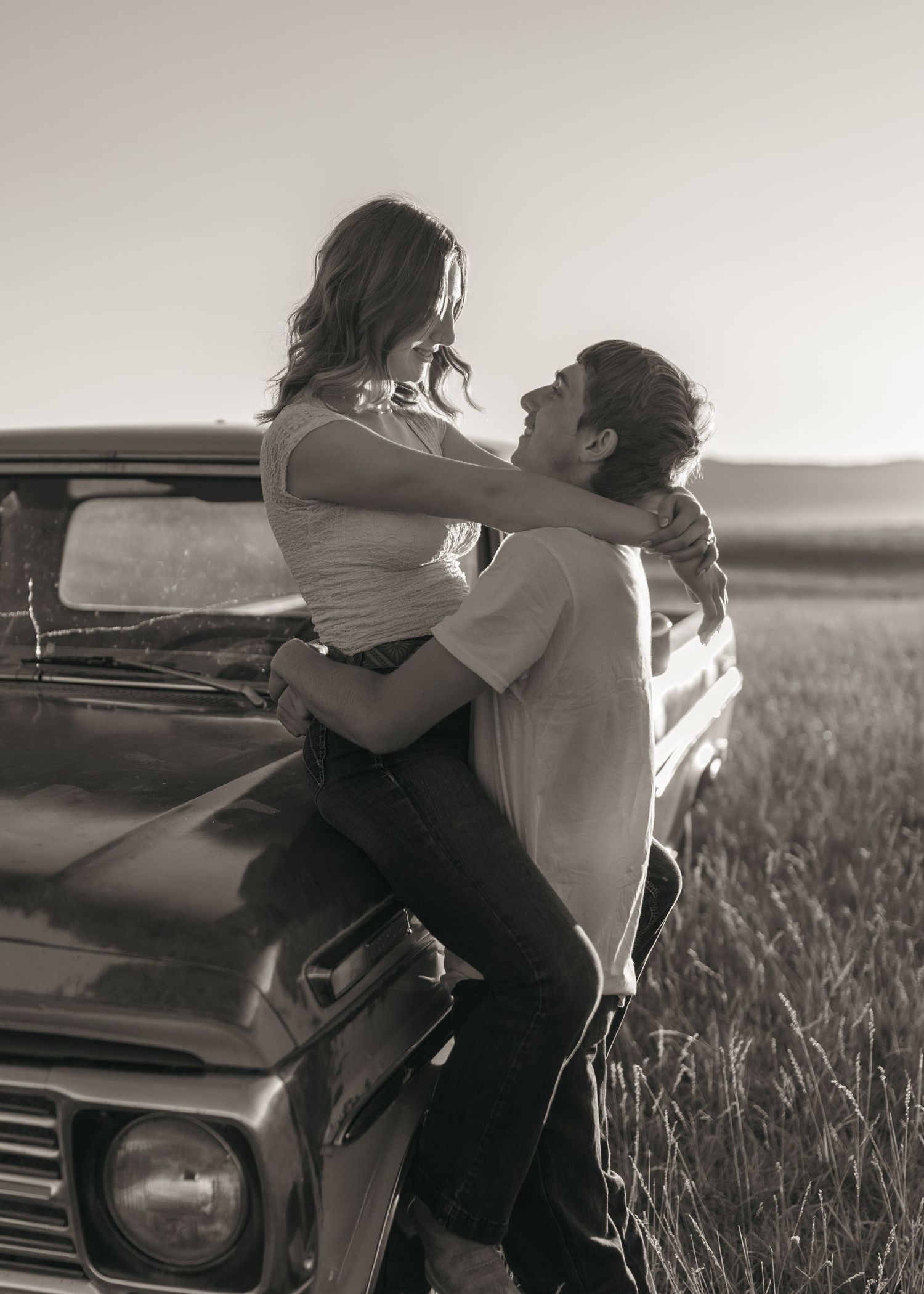 Two figures interact playfully near a classic truck in a rural field setting captured in monochrome.