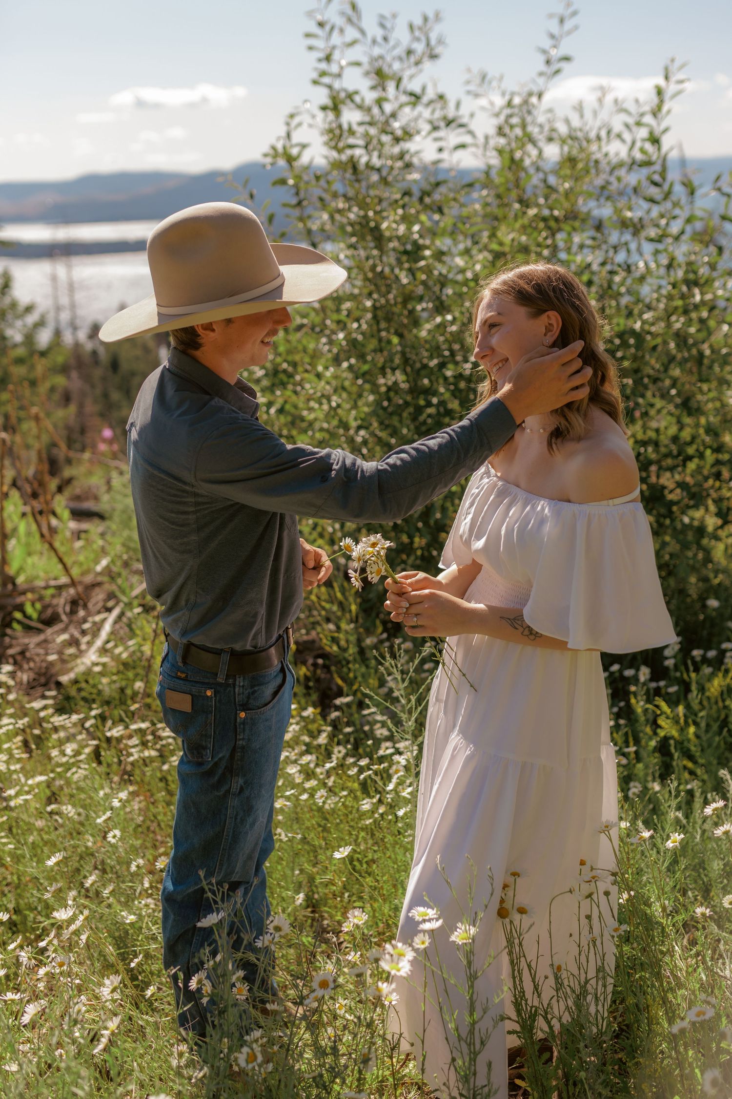 A romantic couple embraces in a field of wildflowers against a scenic mountain backdrop.