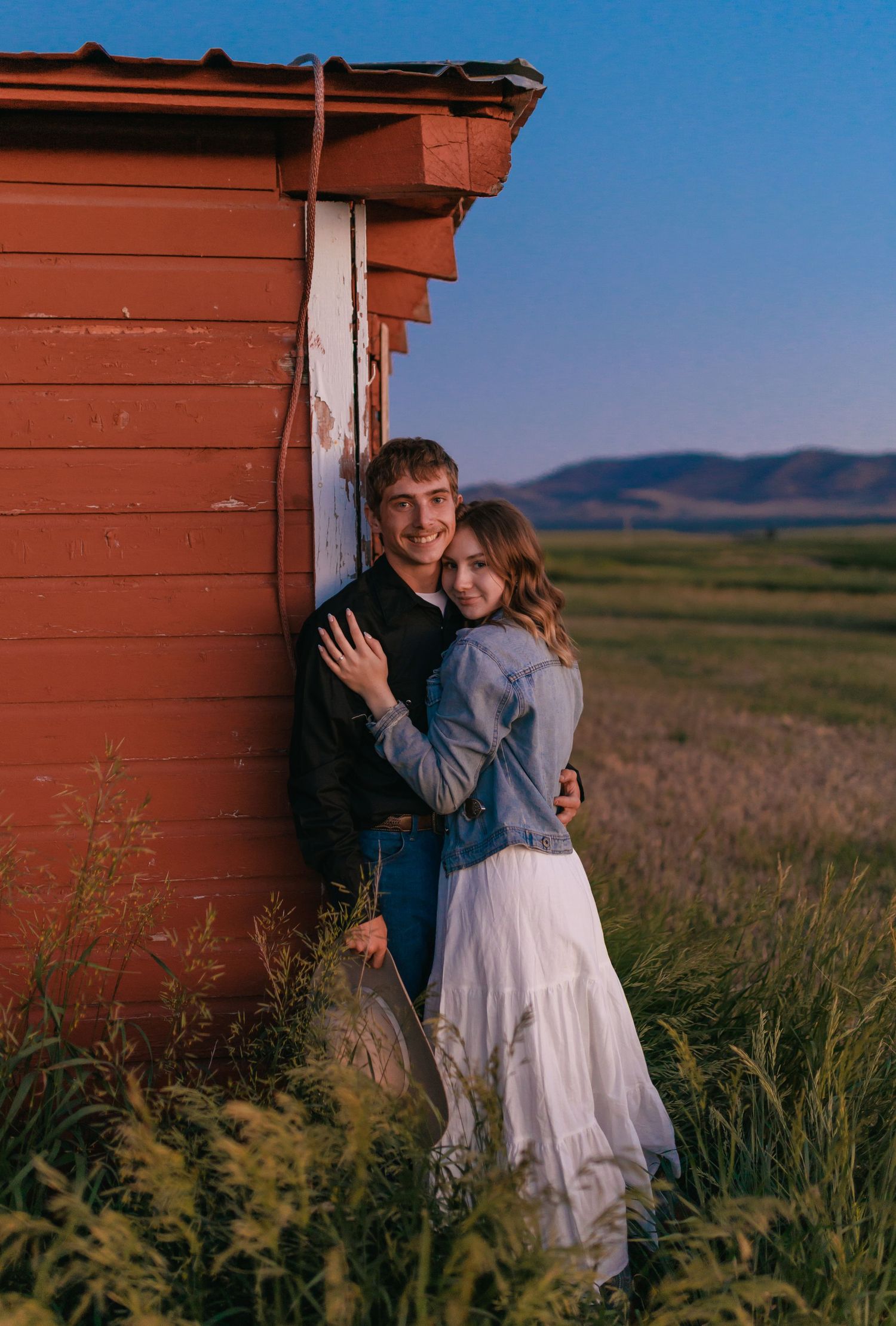 A couple shares an embrace against a rustic red barn in golden evening light.
