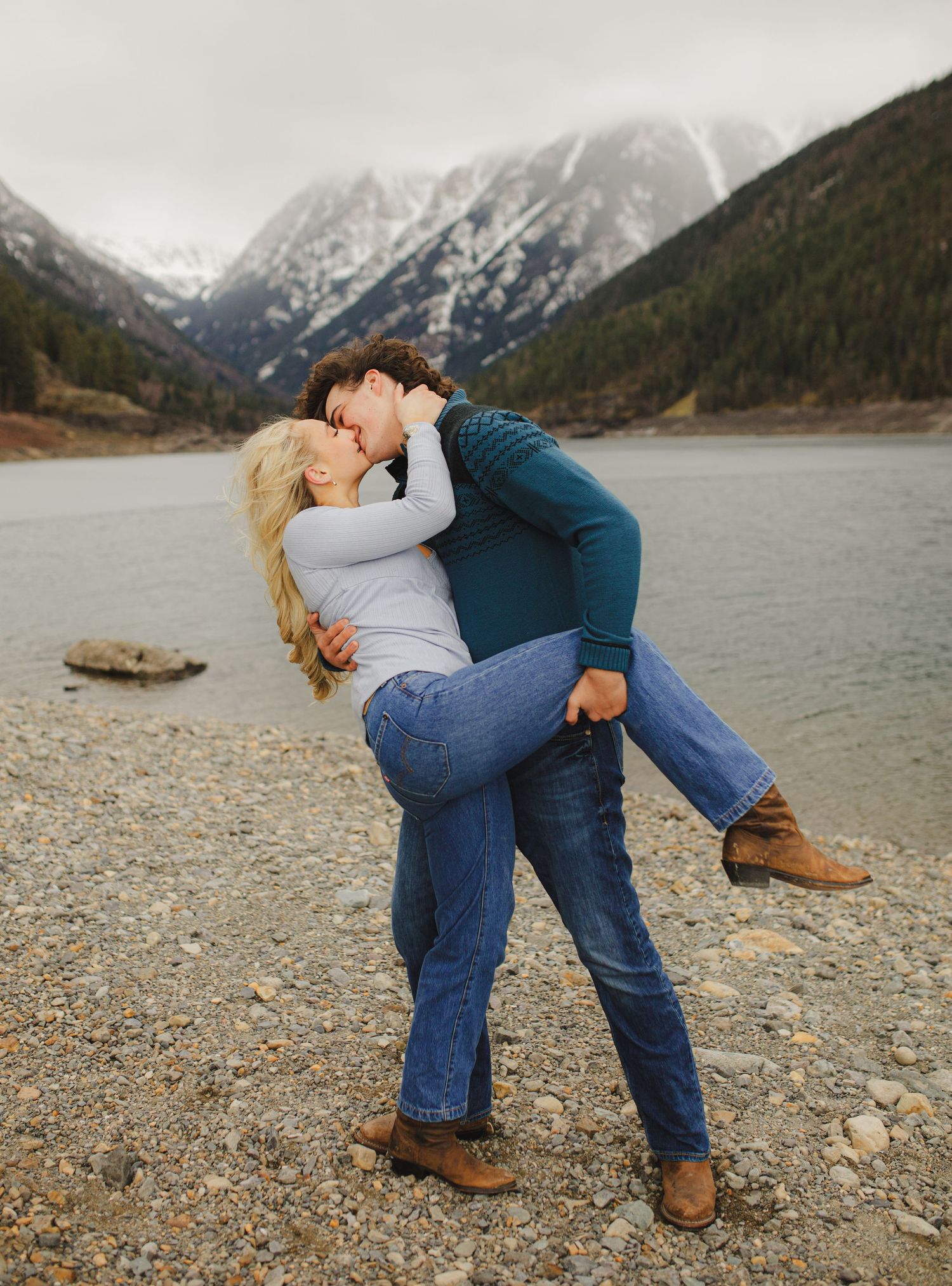 A couple shares a playful dip kiss pose against a backdrop of snowy mountains.