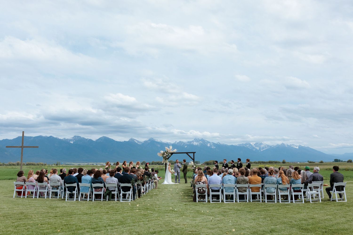 Montana Outdoor wedding ceremony with mission mountain backdrop, guests seated in white chairs facing couple at wooden arch.