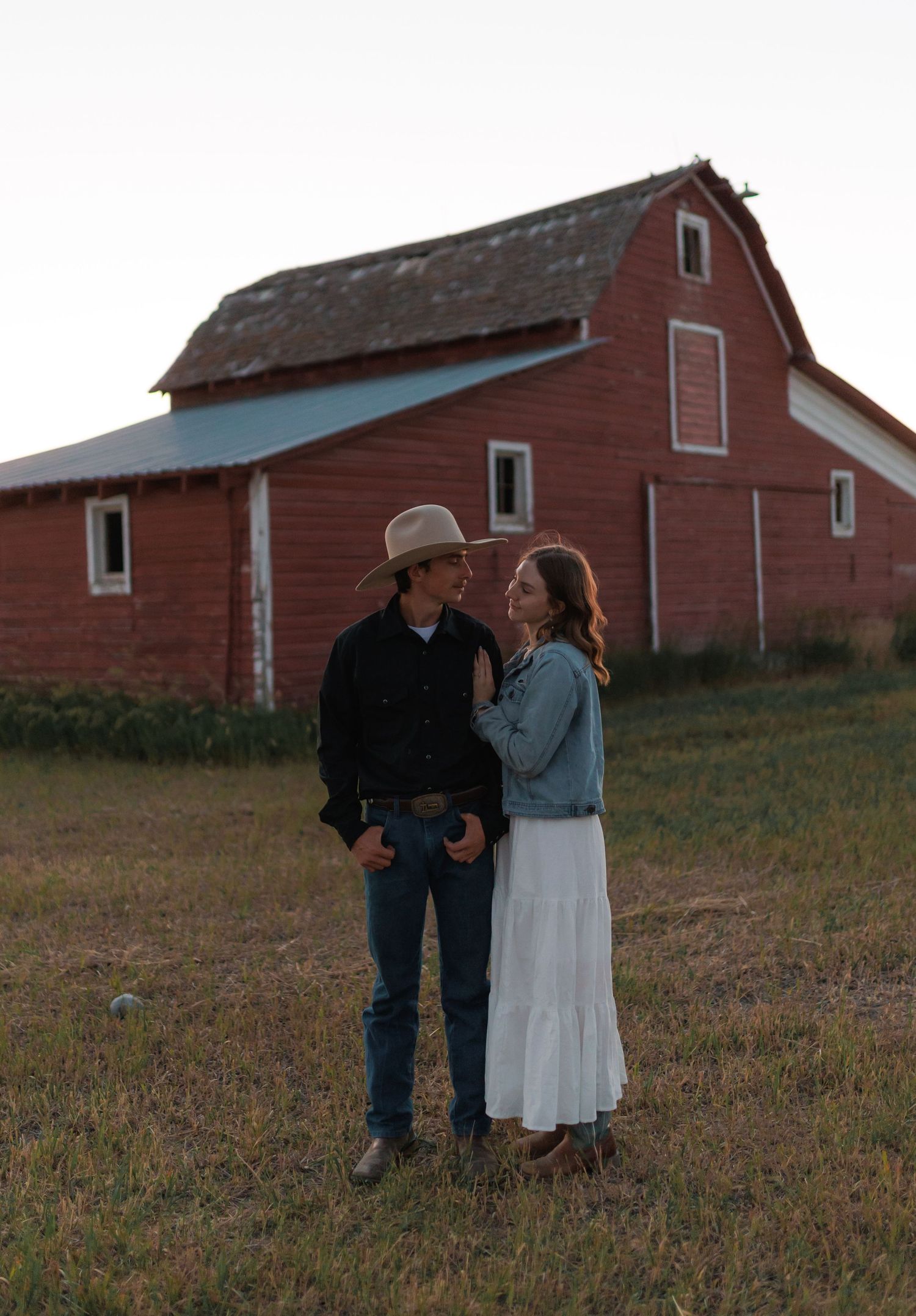 Two people share tender moments beside weathered red farm buildings at dusk.