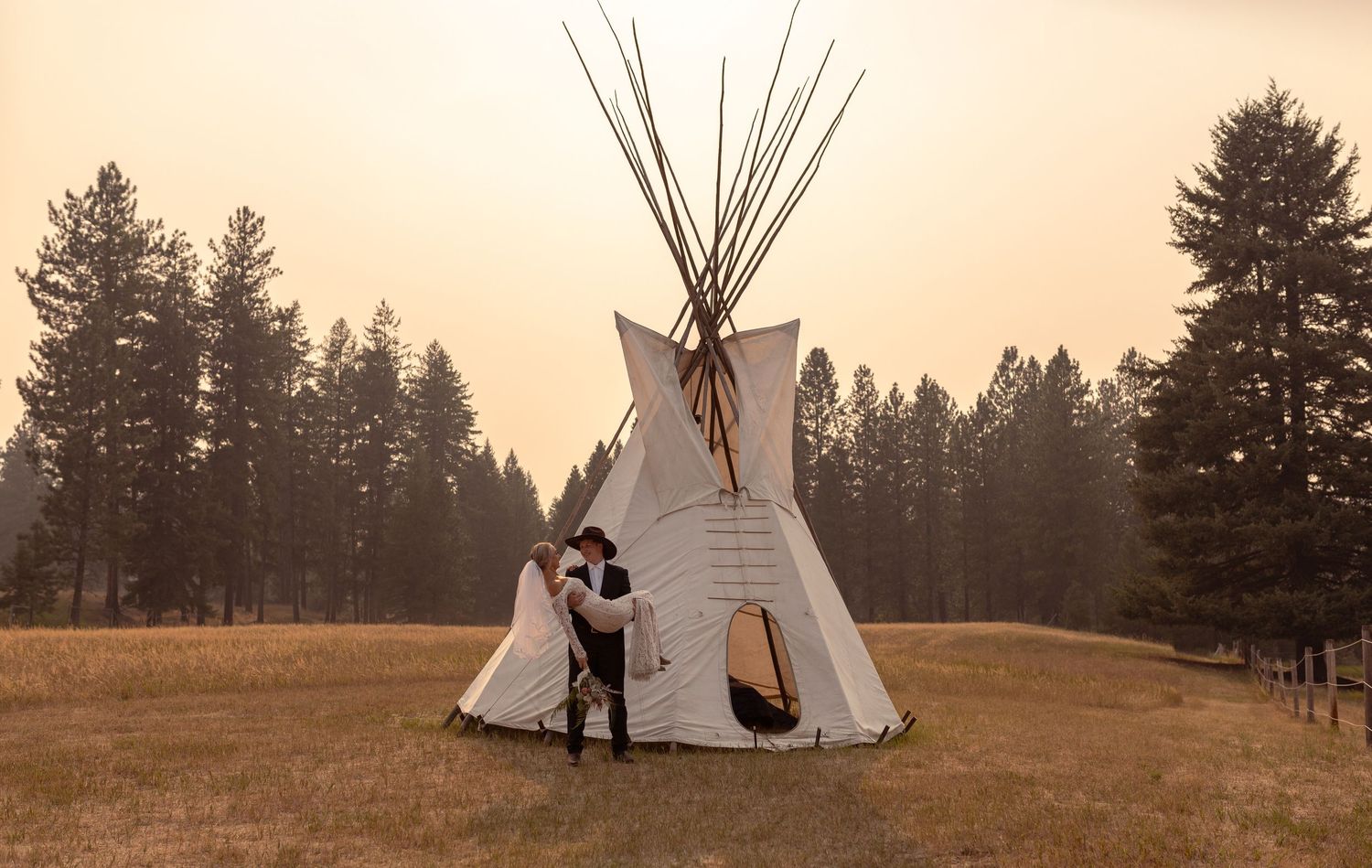 A traditional Native American teepee stands in a golden field with evergreen forest in the background at sunset.