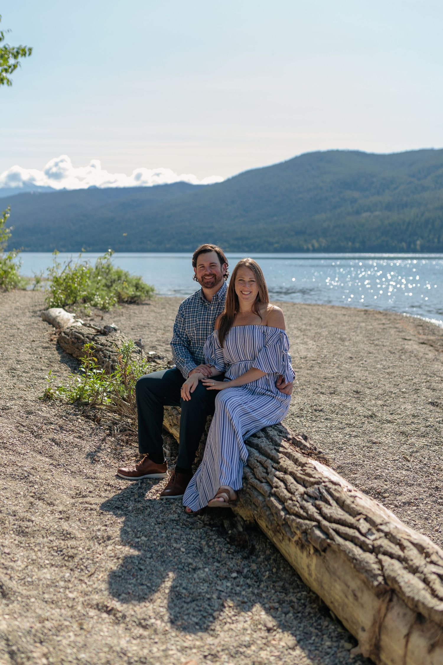 Couple sitting on driftwood log at scenic mountain lake shore on a sunny day.