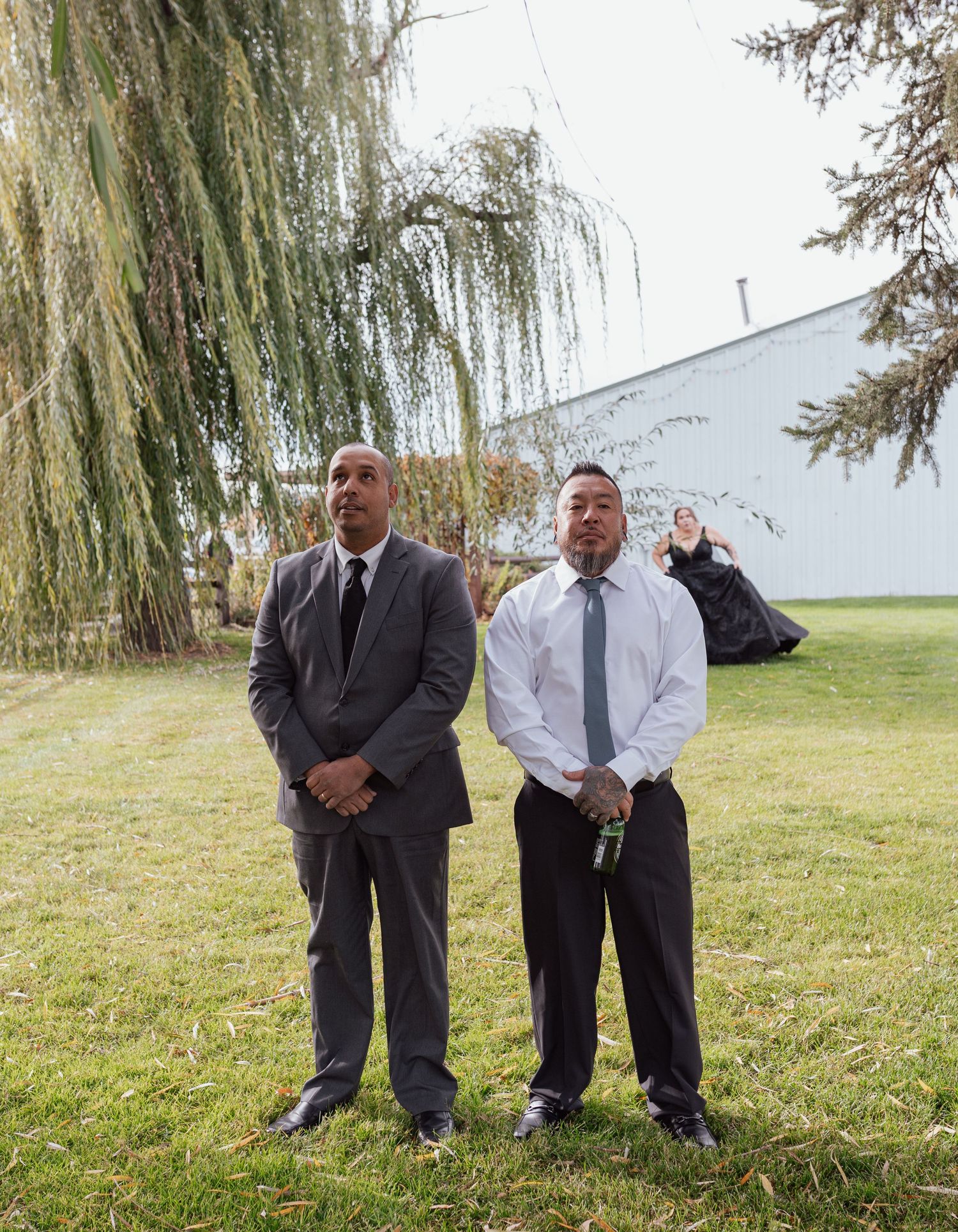 Two formally dressed people stand under a weeping willow tree at an outdoor event.