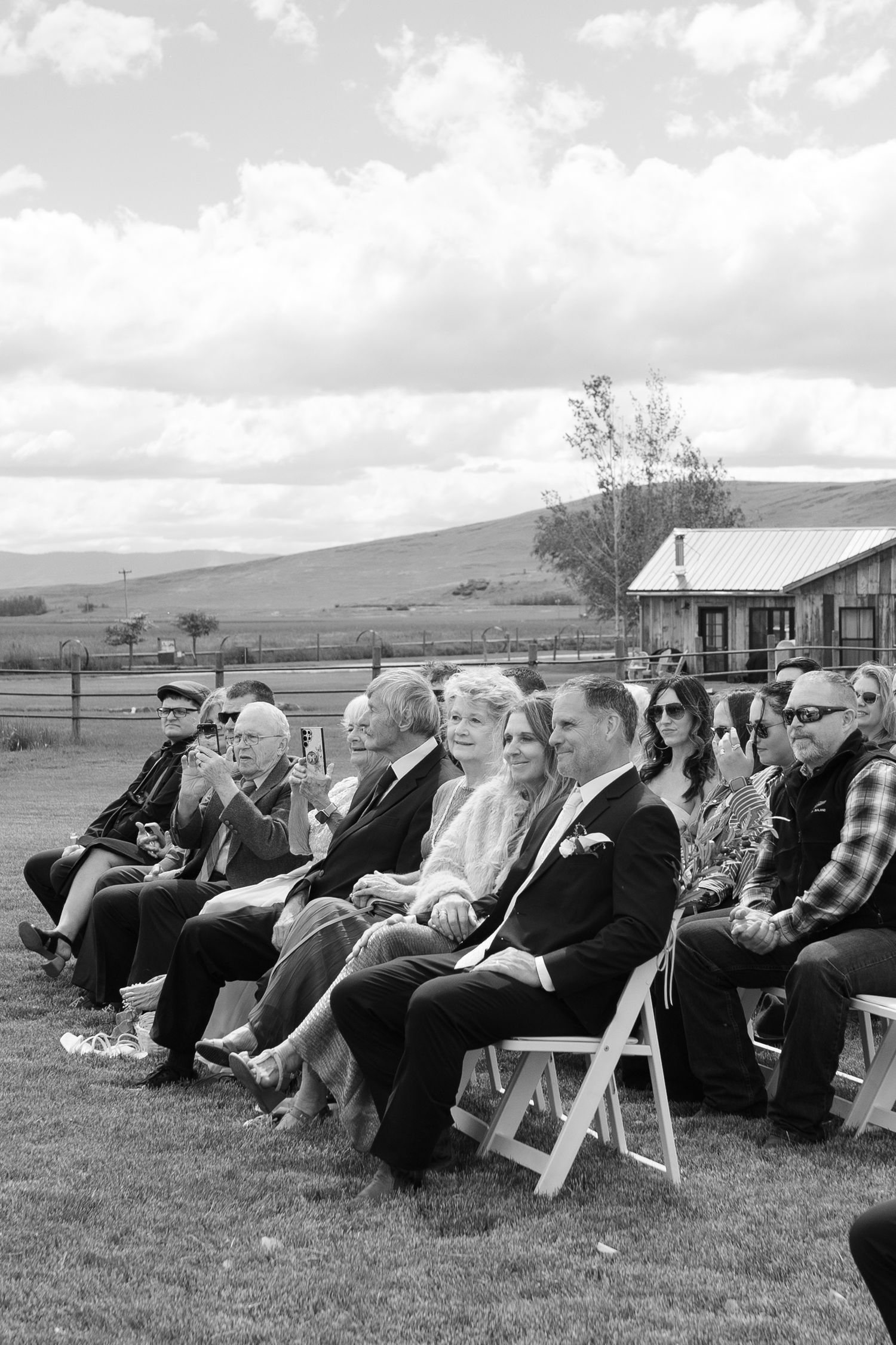 Black and white photo of parents and grandma seated in white chairs outdoors at a rustic ranch ceremony in Montana with mountain views