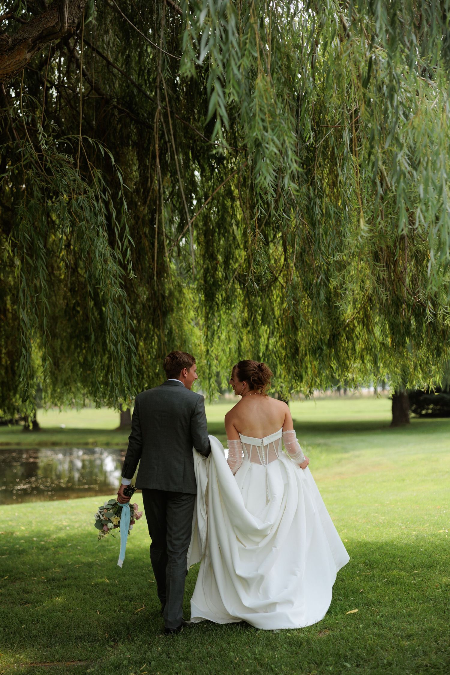 Bride and groom walking hand in hand under a weeping willow tree on lush green grass on their wedding day in Montana.