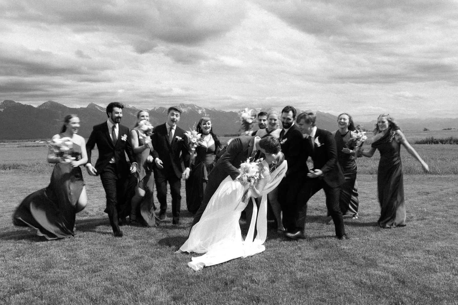 Black and white wedding party photo with bride and groom kissing while bridal party celebrate outdoors with Mission mountains in background.