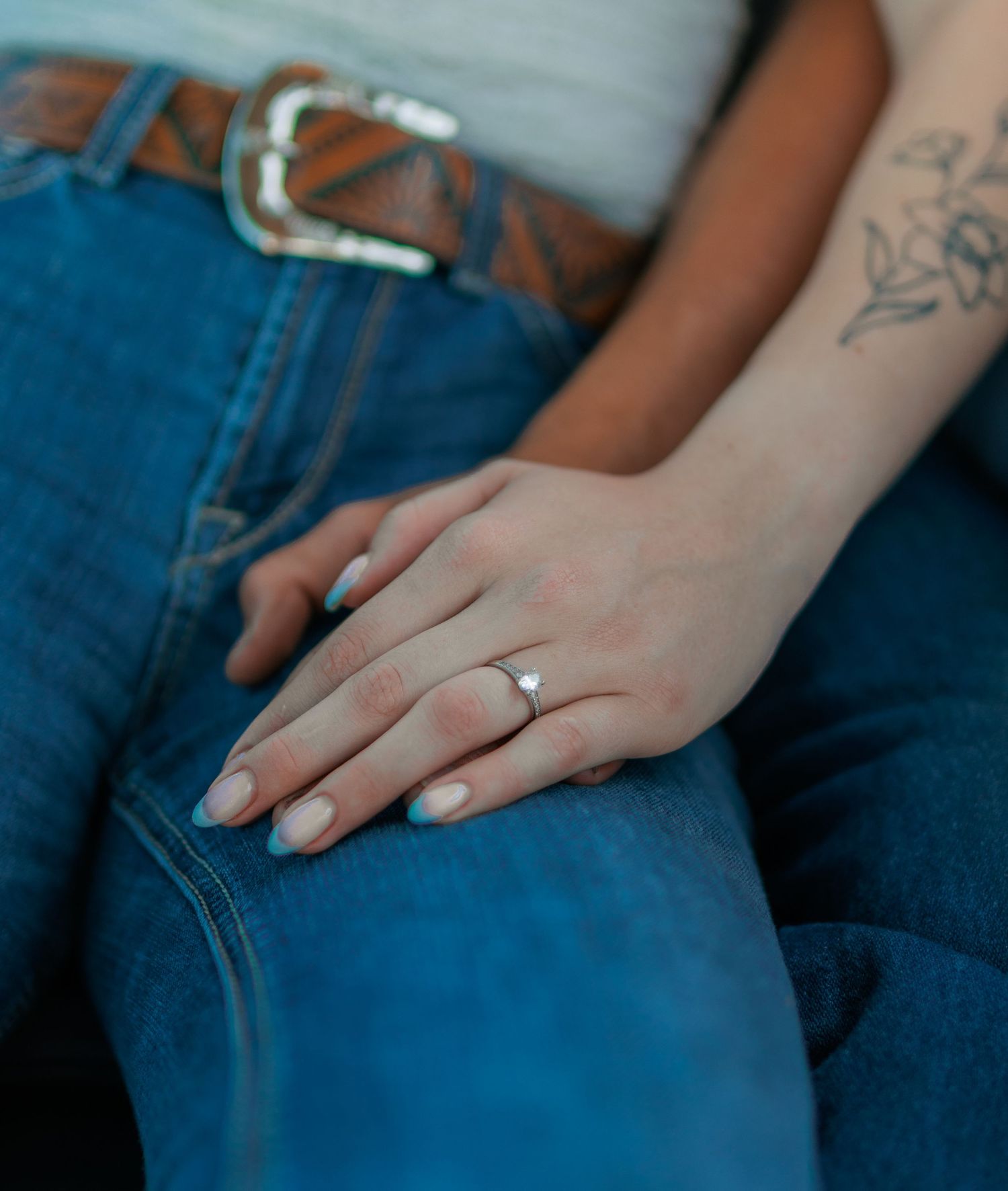 Close-up of hand wearing engagement ring resting on denim jeans with leather belt detail.