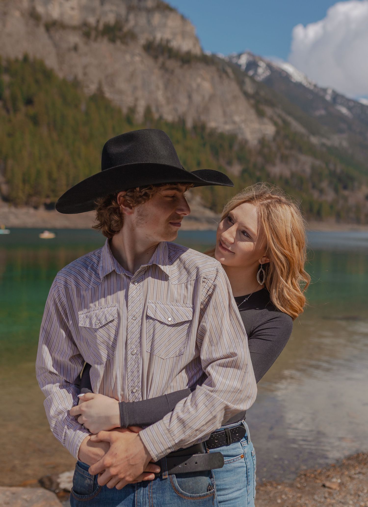 A couple in western attire shares a tender moment by a mountain lake.