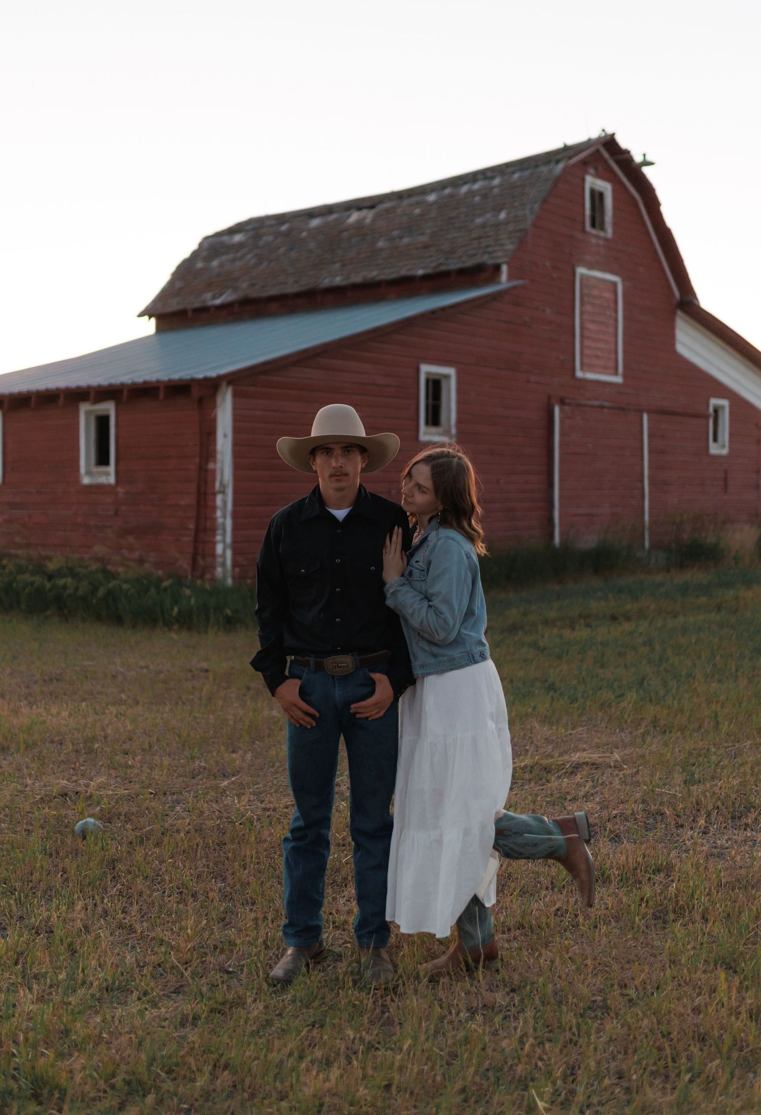 A couple stands together in front of a rustic red barn in a rural field at dusk.