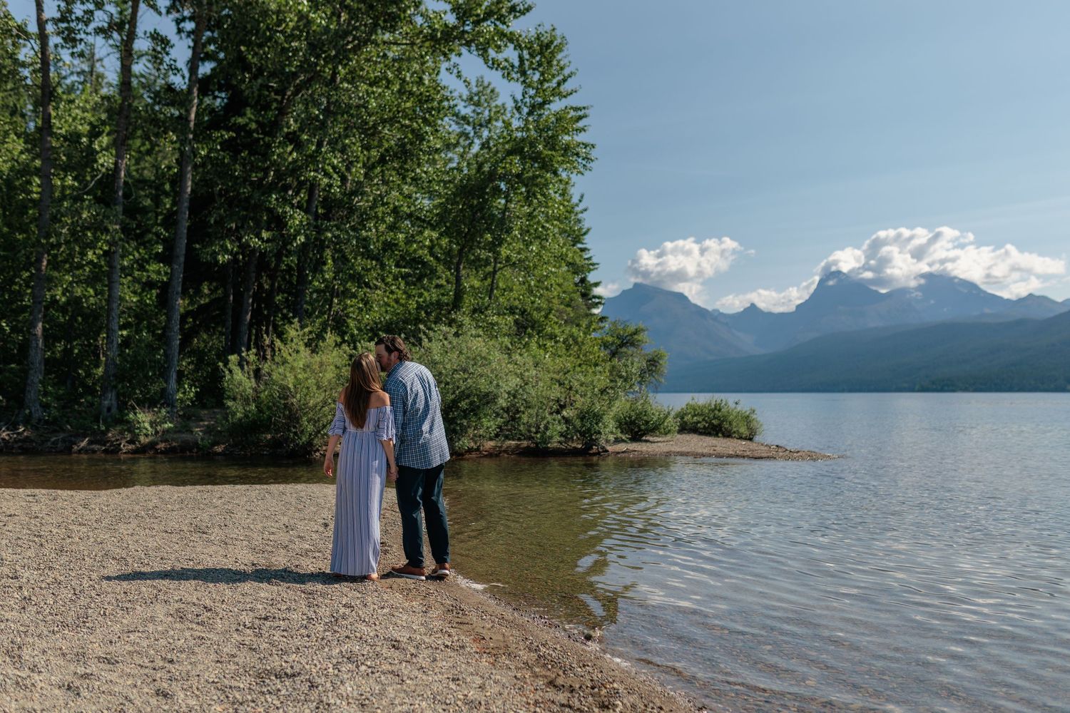 Couple enjoys a summer stroll along the scenic mountain lake shoreline with lush trees and distant peaks.