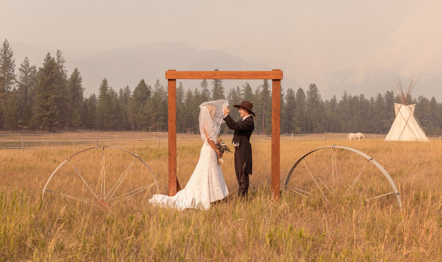 A couple in Montana embraces in a rustic wooden arch frame on a golden meadow with a teepee, white horse, and forest in the background.