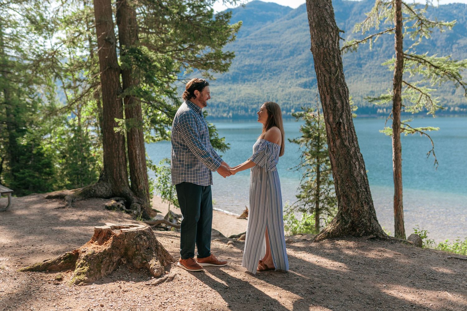 A romantic moment on a lakeside trail with pine trees and mountains in the background.