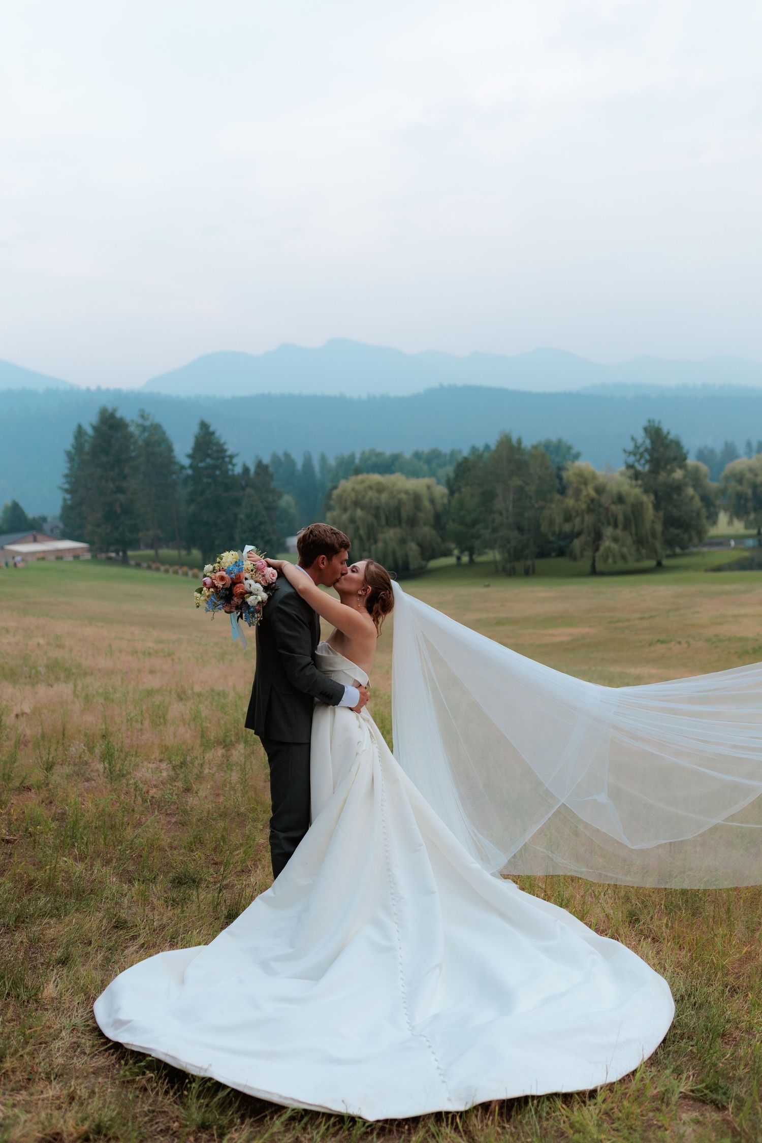 Montana Bride and groom kissing in a mountain meadow, her veil flowing in the wind, holding a colorful bouquet with wildfire smoke at sunset