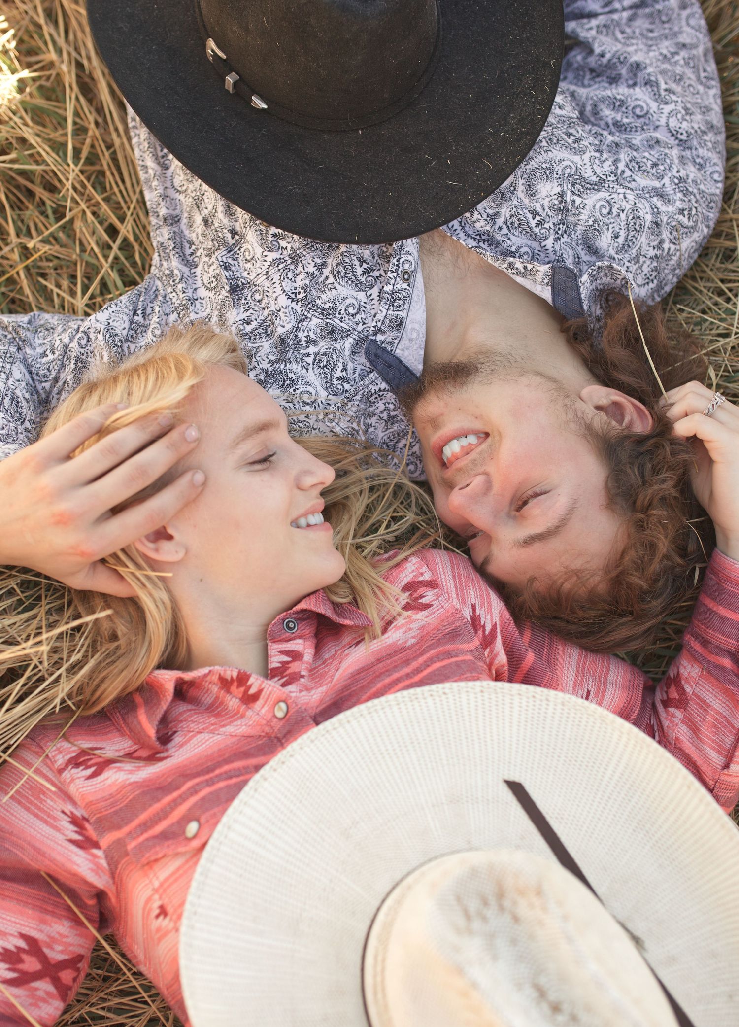Couple lying in hay wearing western hats shares playful laughter.