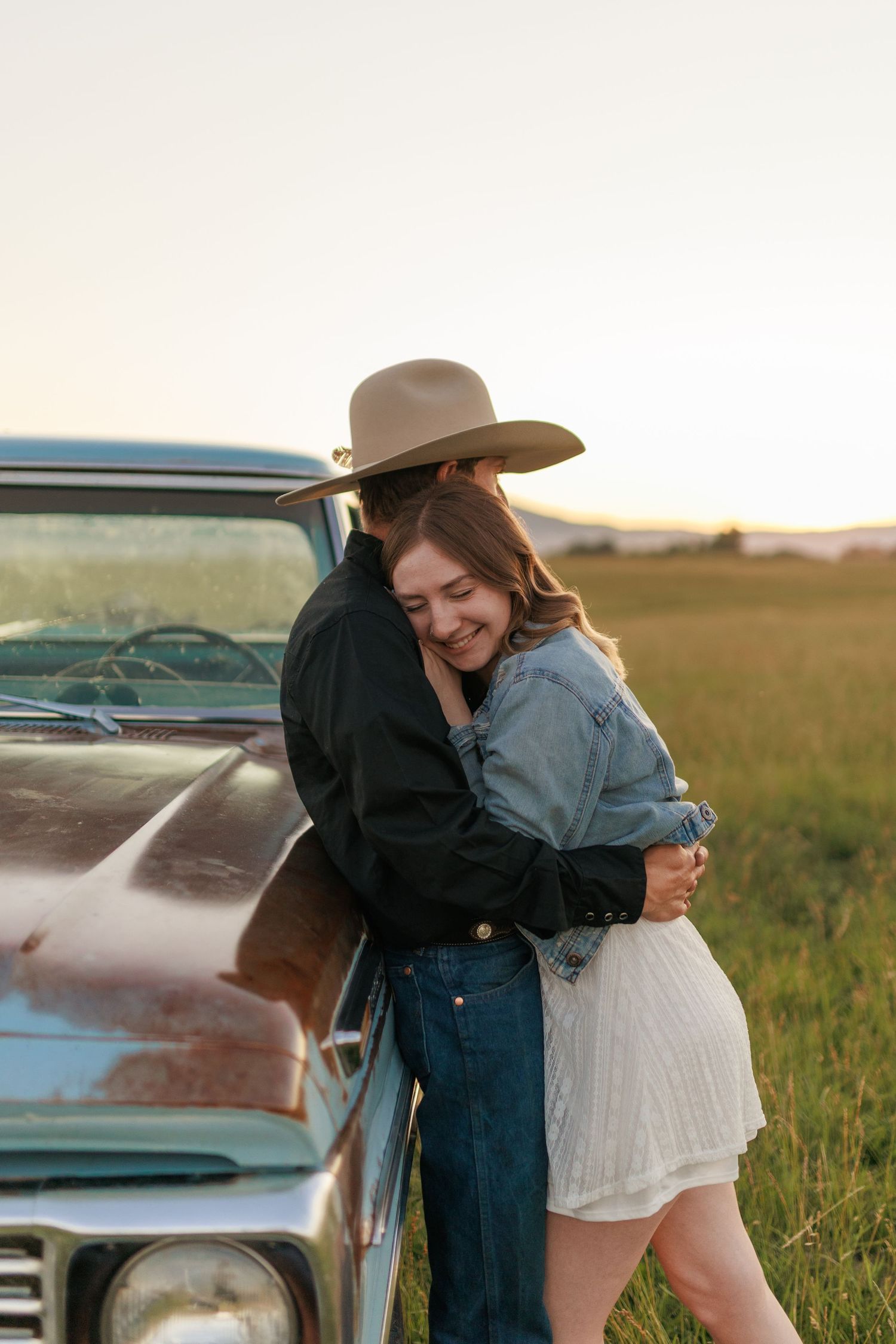 Joyful couple embracing near vintage car in prairie field during golden hour.