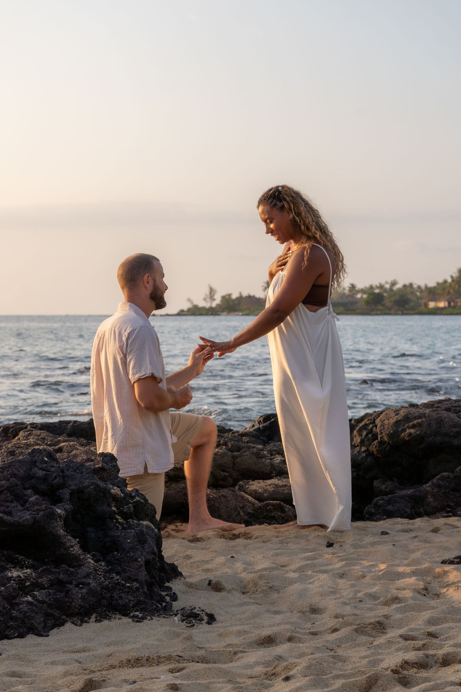 Man proposes to woman on sandy beach at sunset, both in white outfits, with ocean and palm trees in background.